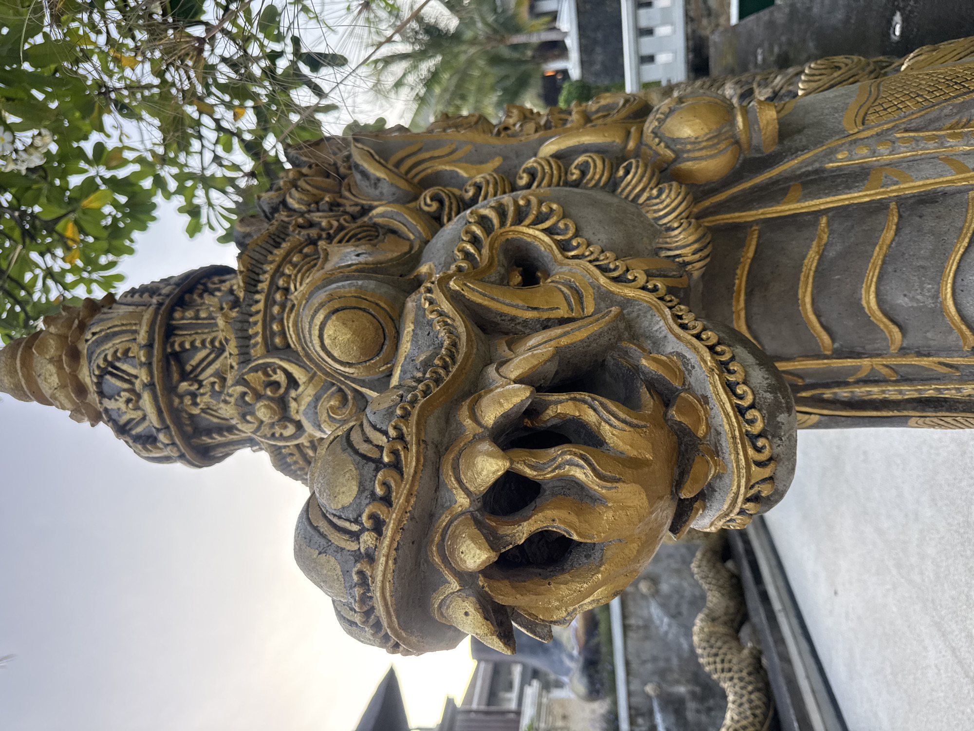 The image shows a close-up, low-angle view of a stone statue with golden embellishments, likely of Balinese origin. The statue appears to be a mythological creature or deity, featuring a detailed face with prominent eyes, ornate headgear, and decorative carvings. The statue's surface exhibits a weathered appearance, suggesting age and exposure to the elements. The setting includes some foliage and architecture in the background, hinting at an outdoor location. Based on the prompt, the statue is located in Kuta Selatan, Indonesia.