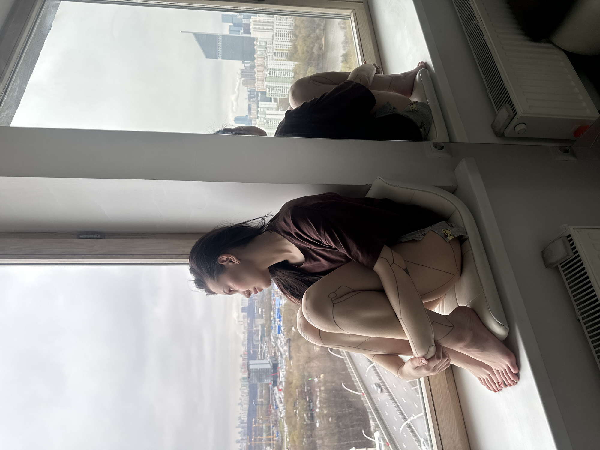 A young woman sits on a windowsill, looking out the window. She is wearing a brown shirt and shorts. Her legs are covered in what appears to be tattoos. The window offers a view of a city, specifically the city of Khimki, Russia, as discernible in the background, with buildings and a highway visible. Part of her is reflecting in what looks like a mirror on the wall. A radiator is on the right side of the frame.