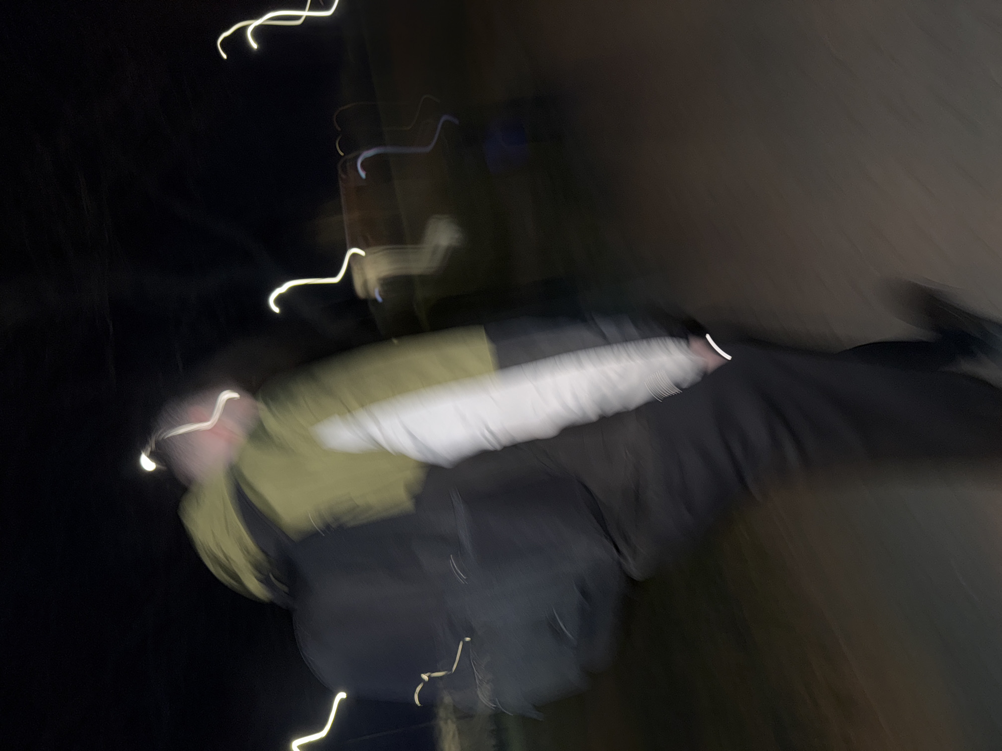 The image shows a blurred figure of a person wearing a jacket with green, white, and black panels. The person appears to be walking or in motion. The environment is dark, with streaks of light, possibly from street lamps or other light sources. The location is unspecified and it is not possible to deduce the location to Łódź, Poland, from the image.