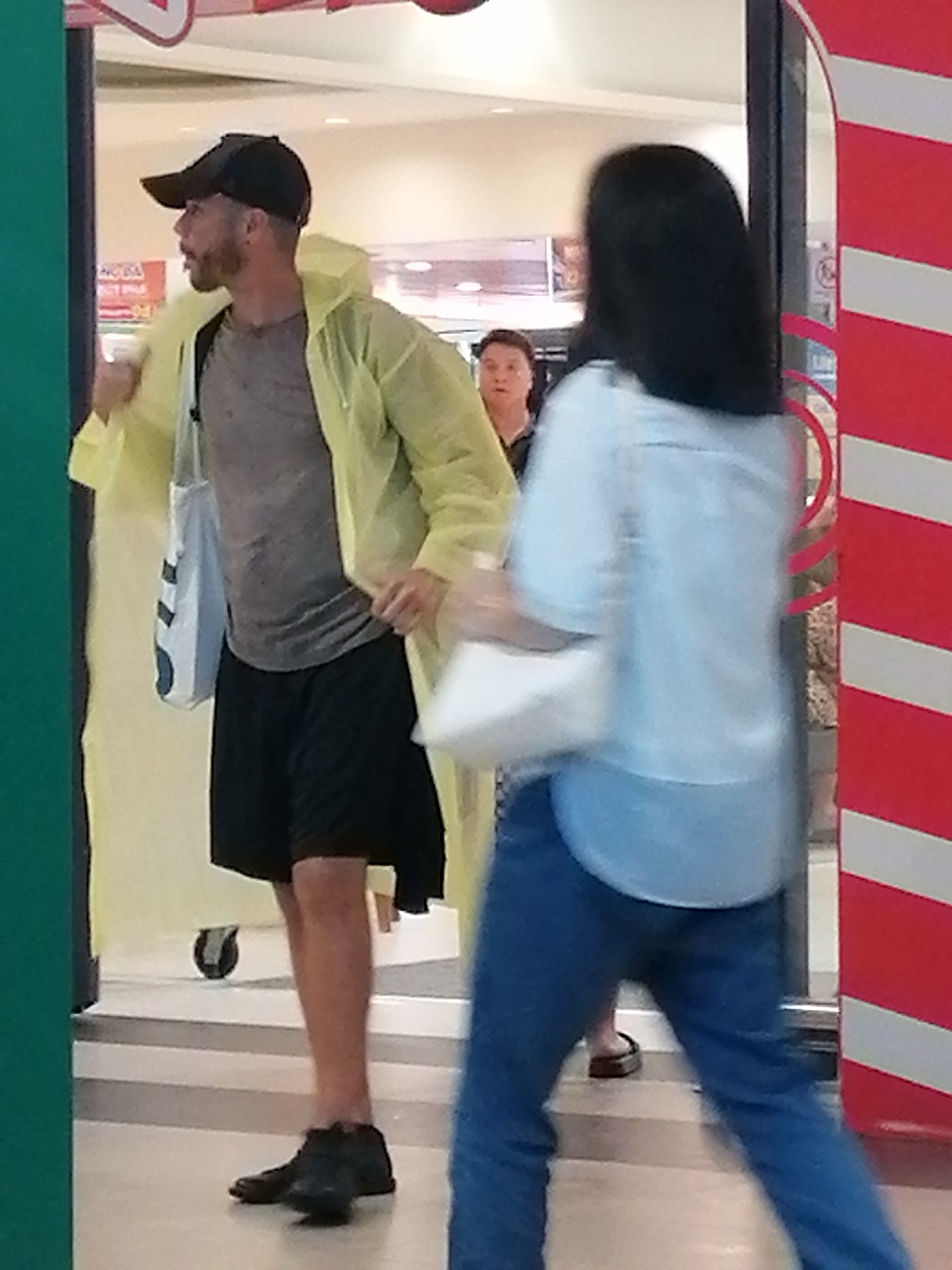 A man wearing a yellow hooded poncho, a grey t-shirt, and black shorts stands on a tiled floor. He has a black baseball cap on and a beard. A white tote bag with black straps hangs over his left shoulder. He is looking to his left. Behind him, a woman with dark hair and a light blue top and blue jeans walks away from the camera, her body blurred. Another woman with short dark hair is visible between the man and the woman in blue. The scene appears to be indoors, possibly in a public space like a shopping mall or an arcade, with a red and white striped pillar on the right side of the image. The context of Da Lat, Vietnam is not apparent from the visual information in the image.