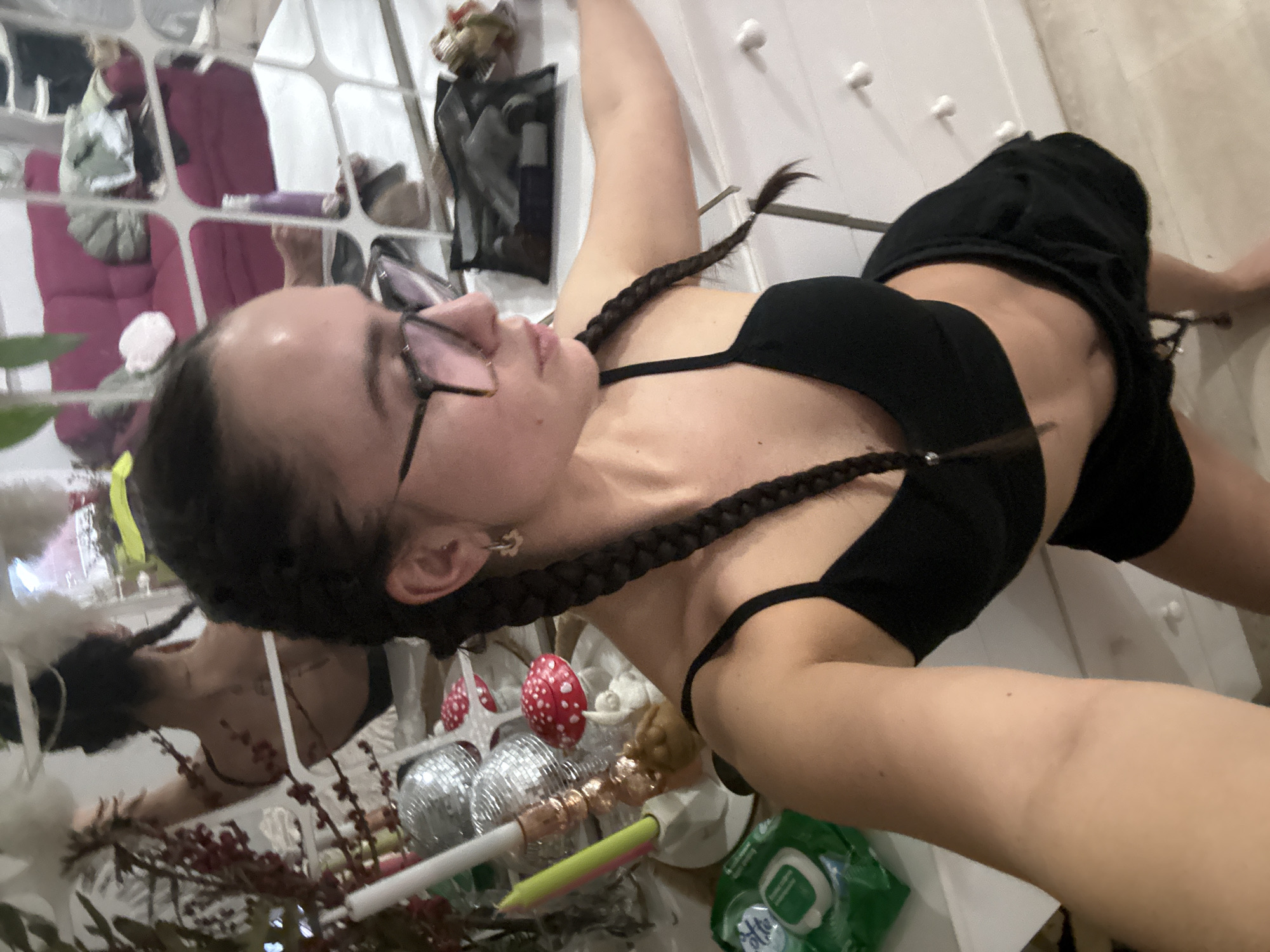 The image features a young woman in an indoor setting, likely taking a selfie from a low angle. She is lying down or reclining, looking upwards. Her dark hair is styled in two long braids, and she wears dark-framed glasses, a black bra top, and black shorts. Her body is slender, and her expression appears relaxed and casual.
The background includes a mirror or window with a grid pattern, reflecting various items from the room, such as a reddish-purple armchair or cushion, clothing, and other decorative elements. To the right, a white wall or piece of furniture with white knobs is visible.
In the foreground and scattered around, there are several decorative objects: red berry-like branches, two silver disco ball ornaments, and a red and white spotted mushroom ornament. A white candle with yellow and pink sections is also present, along with a green packet, which appears to be wet wipes, with partially visible text that reads "Wet Wipes".
The overall scene suggests a personal and relaxed atmosphere, possibly in a bedroom or living space in Krasnoyarsk, Russia. The time of day is indeterminate, but the lighting suggests an indoor setting.