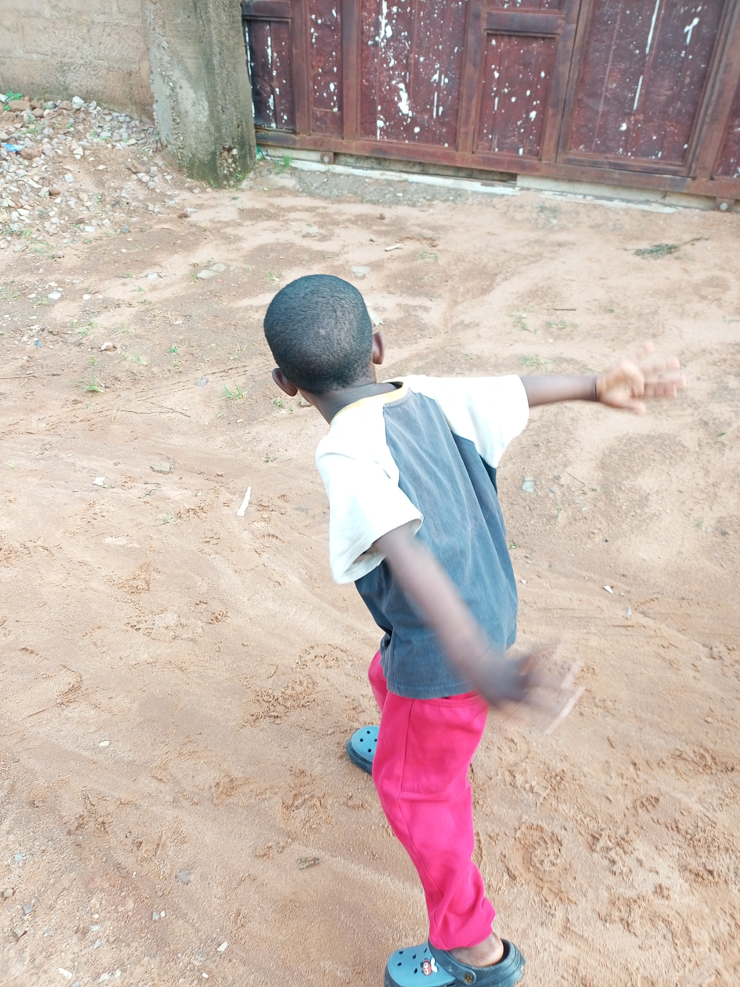 A young boy with dark skin and short, dark hair is standing on sandy ground. He is wearing a t-shirt with white sleeves and a gray torso, and bright pink pants. On his feet are blue, open-toed shoes. His arms are outstretched, and his hands are blurred with motion, suggesting he is actively engaged in an activity. The background consists of a rough, concrete wall on the left, with a pile of small rocks and debris next to it. To the right, there is a large, weathered wooden gate with a dark reddish-brown color and white splatters. The ground is a mix of sand and some sparse, dry vegetation. The scene appears to be outdoors in what could be a residential or outdoor area in Tempure, Nigeria.