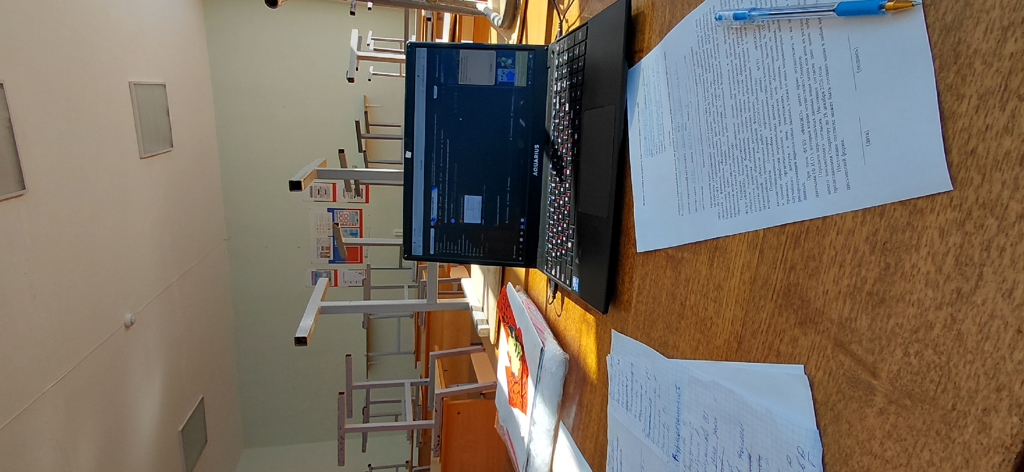The image depicts a classroom scene in Svetlogorsk, Russia. The focus is on a wooden desk in the foreground with various items laid out. A laptop, branded AQUARIUS, is open, displaying what appears to be a web page or application with text and some visual elements. To the left of the laptop, there are stacks of papers, some with handwritten notes and others appearing to be printed documents. A blue pen is positioned on the right side of the desk, resting on a printed form with Cyrillic text.
In the background, the classroom is visible with rows of empty student desks and chairs, some stacked. The walls are light-colored, and the ceiling has fluorescent lighting fixtures. The overall impression is one of a quiet, possibly unoccupied classroom, with an active workspace set up at the desk. The sunlight streaming in from a window casts shadows across the desk and papers.