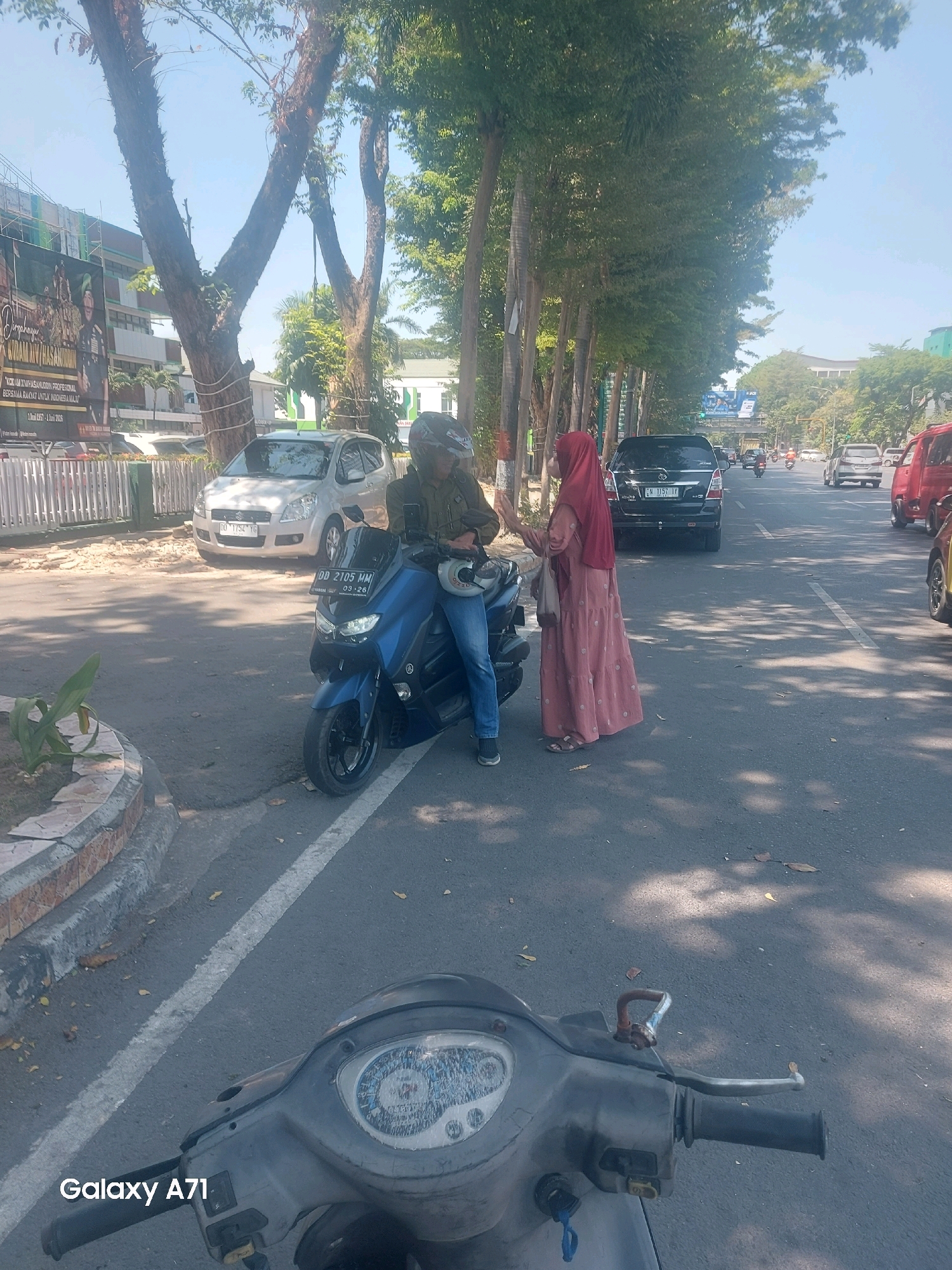 The image shows a street scene in Makassar, Indonesia, with several vehicles and people. In the foreground, the dashboard and handlebars of a scooter are visible, indicating the photo was taken from the perspective of someone riding or parked on a scooter. In the middle ground, a blue Yamaha NMAX scooter is parked on the right side of the road, with a rider wearing a green jacket and helmet. A woman in a pink dress and red hijab is standing next to the scooter, facing the rider and gesturing with her hands. A silver car is parked behind the scooter. Further down the street, other vehicles, including cars and motorcycles, are moving. Trees line the left side of the road, with a building and a large banner visible in the background. The overall environment appears to be an urban street on a sunny day.