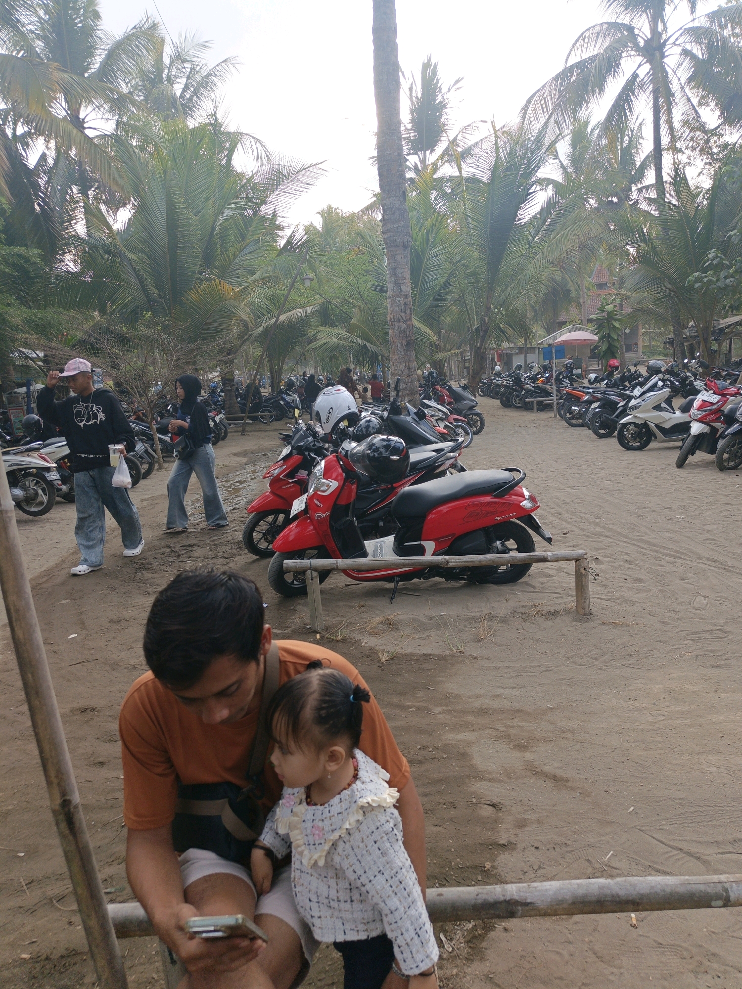In Tambakmulyo, Indonesia, a man and a young child are seated on a wooden railing in the foreground, with the man holding a mobile phone. Behind them, a sandy area is filled with parked motorbikes. A few people are visible walking amongst the motorbikes. The background features numerous palm trees and a cloudy sky. The scene appears to be a public parking area, likely near a beach or recreational spot.