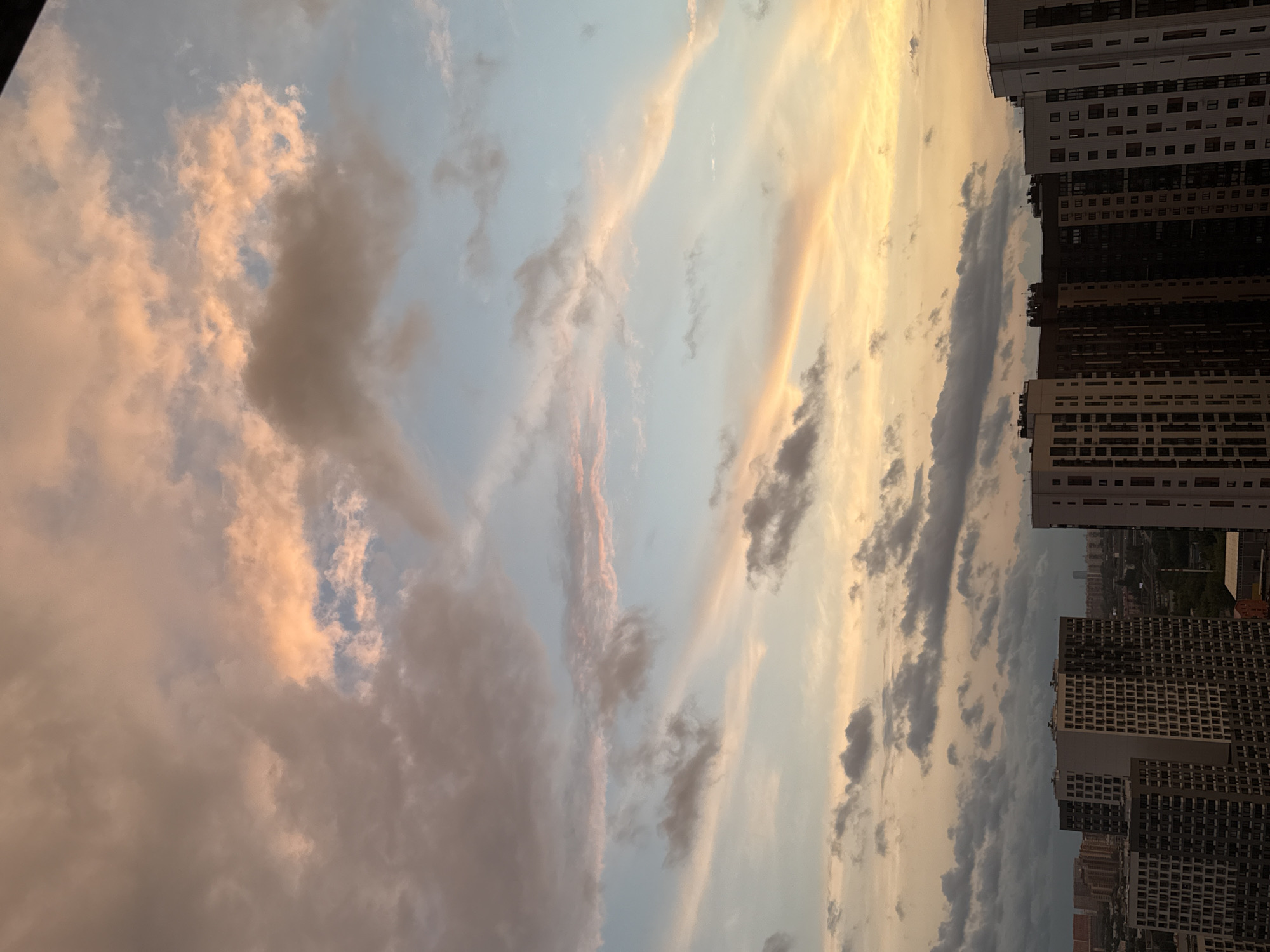 The image depicts a cityscape in Lyubertsy, Russia, at dusk. The sky is filled with clouds tinged with pink and orange hues, indicative of sunset. In the foreground, a cluster of modern high-rise apartment buildings dominates the lower portion of the frame. These buildings are characterized by their repetitive window patterns and varying heights, suggesting a relatively new residential development. The urban environment extends into the background, with more buildings and a distant cityscape visible beneath the dramatic sky. No people or specific actions are discernible. The overall impression is one of an urban evening, with the natural beauty of the sunset contrasting with the man-made structures.