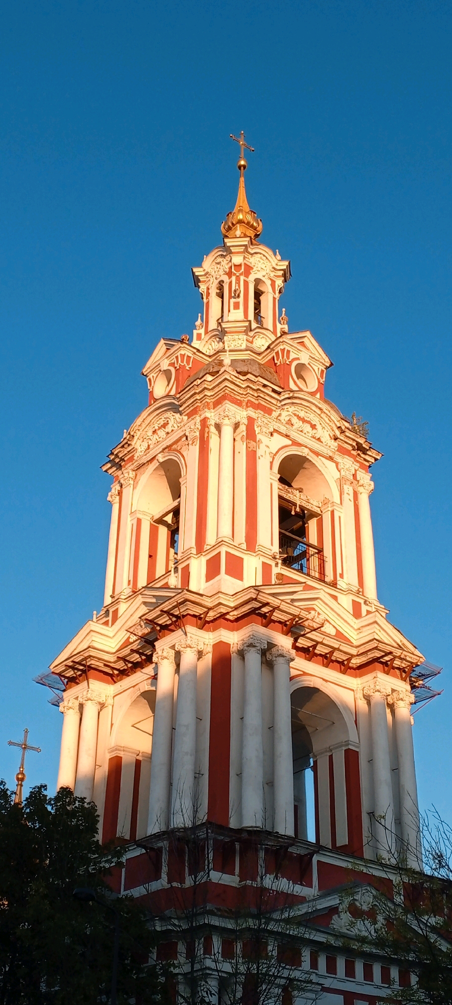 The image shows the bell tower of the Epiphany Monastery in Elokhovo, Moscow, Russia. The bell tower is painted in alternating stripes of red and white and features a golden dome topped with a cross. It is illuminated by the warm light of the setting sun, casting long shadows. The sky is a clear, deep blue. In the foreground, the dark silhouettes of trees and some roofing are visible. A smaller cross on a spire can be seen to the left of the bell tower. The architecture is ornate, with columns, arches, and decorative mouldings visible.