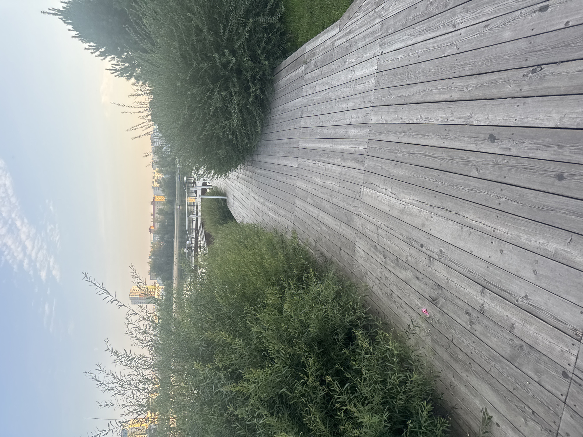 The image depicts a wooden walkway adjacent to a body of water with trees and buildings in the background. The walkway is constructed of weathered gray planks and curves to the left, bordered by dense green bushes on the left and right. In the distance, across the water, a cityscape with several multi-story buildings is visible under a pale sky with scattered clouds. A portion of a white railing and a pier structure can be seen near the water's edge on the left side of the walkway. The overall scene suggests an outdoor recreational area, possibly a park or promenade. The specific location in Ufa, Russia, is not identifiable from the image alone.