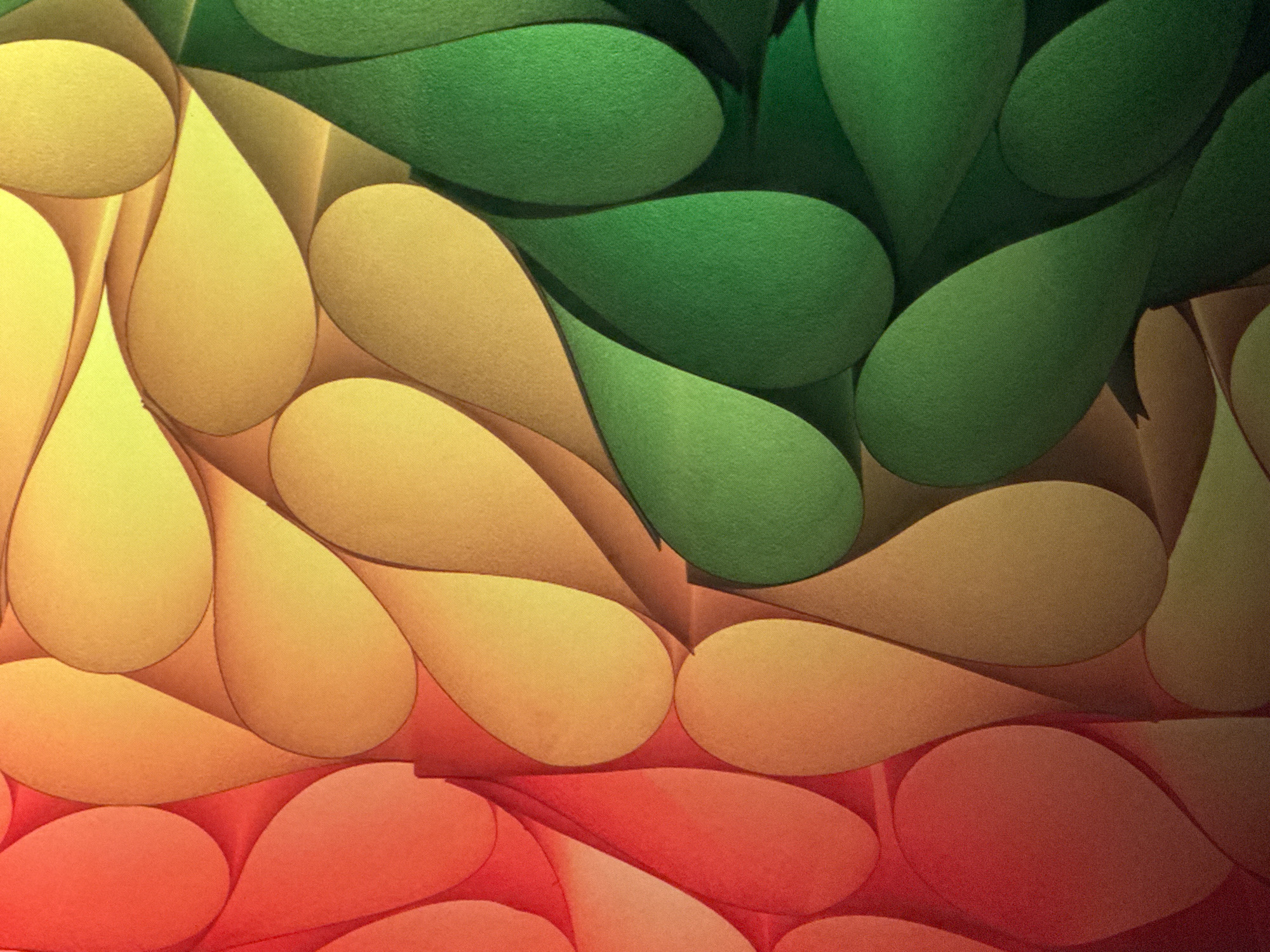 The image is a close-up, abstract photograph of folded paper with a gradient of colors from red to orange, yellow, and green. The paper is arranged in a repeating pattern of teardrop shapes, creating a textured and dimensional effect. There are no people, animals, or recognizable landmarks in the image. The lighting emphasizes the curves and shadows of the folded paper. The image does not depict any location in Saratov, Russia.