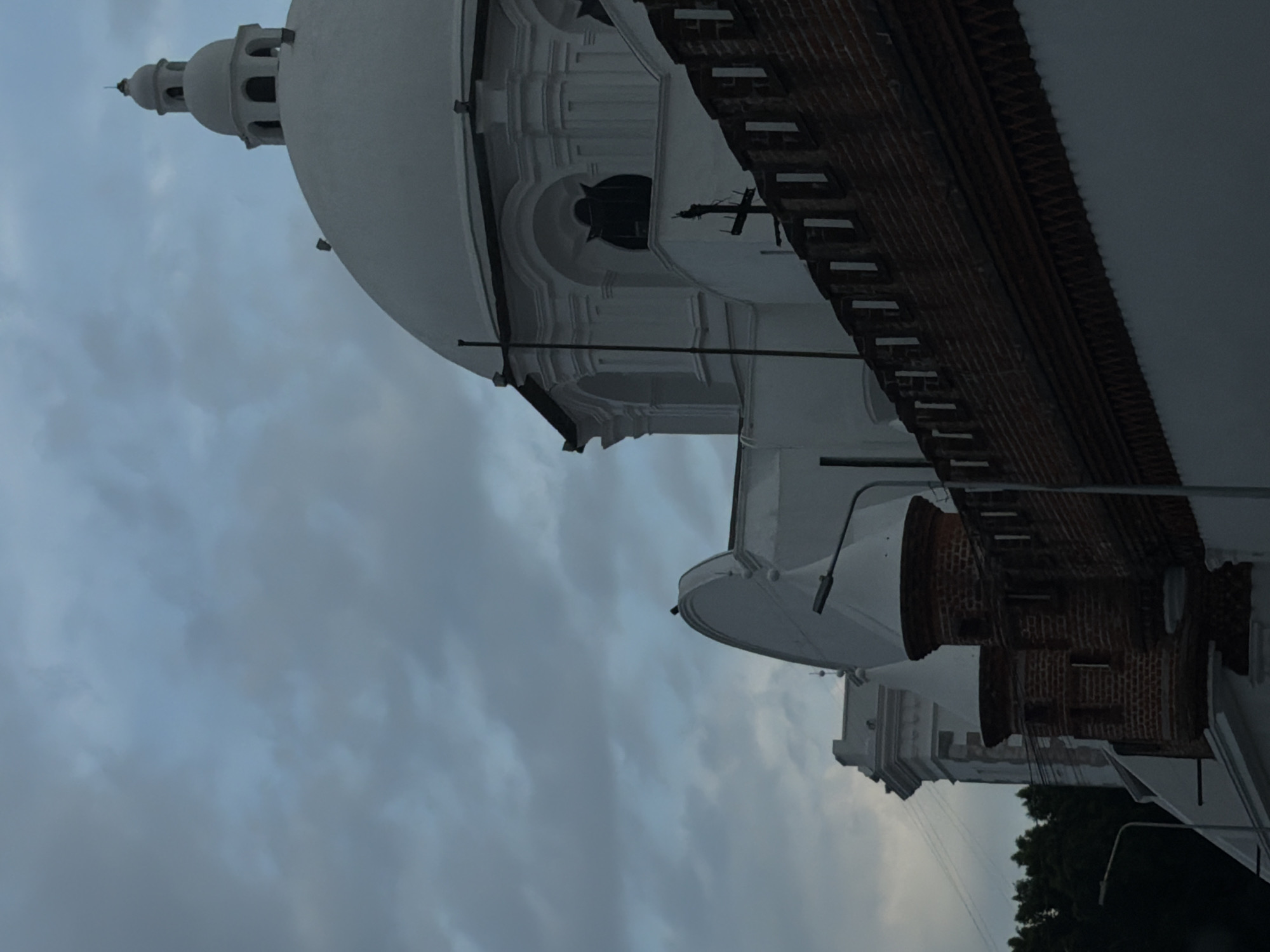 The image depicts a section of a building complex under a cloudy sky. The architecture includes a white dome topped with a smaller tower and windows, a brick facade, and a white tower with a cross. The building appears to be located in Puebla, Mexico, possibly a religious structure or historic site, based on its architectural style. Street lighting fixtures are visible extending from the building.