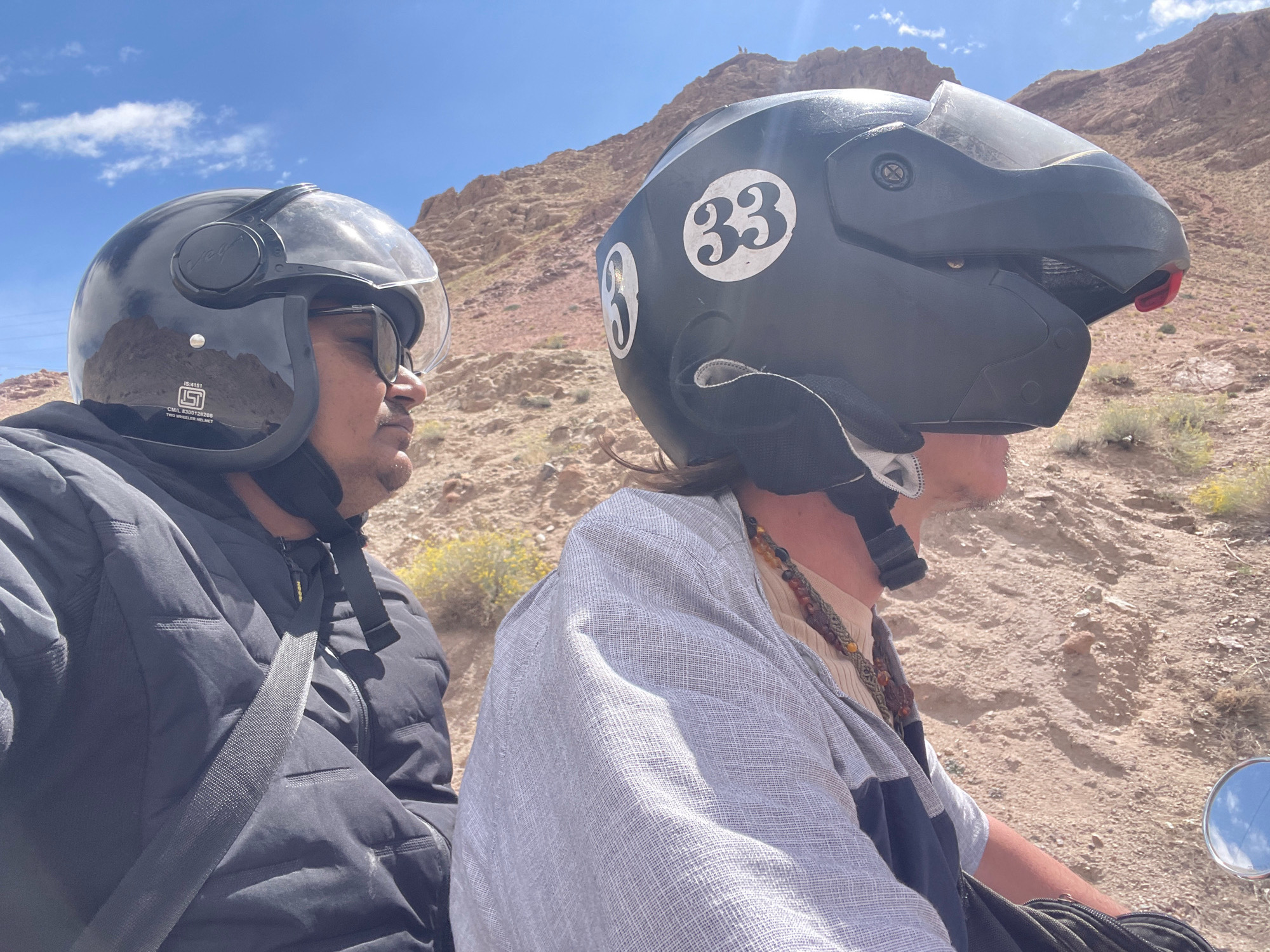 The image shows two people wearing helmets while riding on a motorcycle in a desert environment. The person in the foreground on the left is wearing a black helmet and a black puffer jacket, with sunglasses on. They are looking to the right. The person on the right is wearing a black helmet with a white number '33' decal, and is wearing a light-colored garment over a lighter shirt, with a beaded necklace. They are looking to the right, in the same direction as the motorcycle is presumably traveling. The background consists of dry, rocky hills with sparse vegetation, indicative of a desert or arid region. The sky is blue with some scattered clouds. The location is identified as Arpa, India, which is a region known for its mountainous and arid landscapes. The image captures a moment of travel, likely an adventure or exploration in this unique geographical setting.