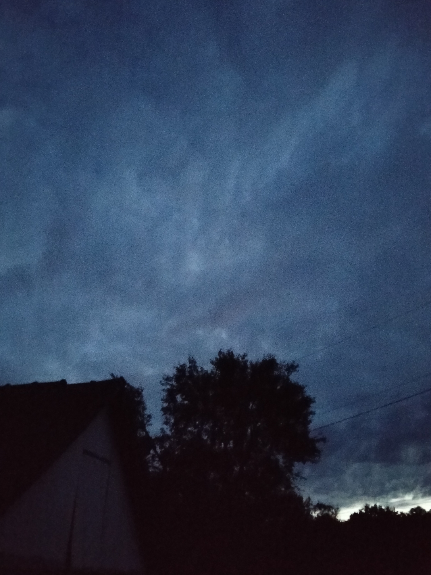 The image depicts a nighttime scene in Ustyvytsia, Ukraine. A dark, triangular structure, likely a building, occupies the lower left portion of the frame. To the right of the structure, a dense tree with dark foliage is silhouetted against the sky. The sky itself is filled with dark, cloudy formations. A faint power line is visible stretching across the upper right quadrant of the image. The overall atmosphere is dim and overcast, suggesting the presence of twilight or a very dark, cloudy night.
