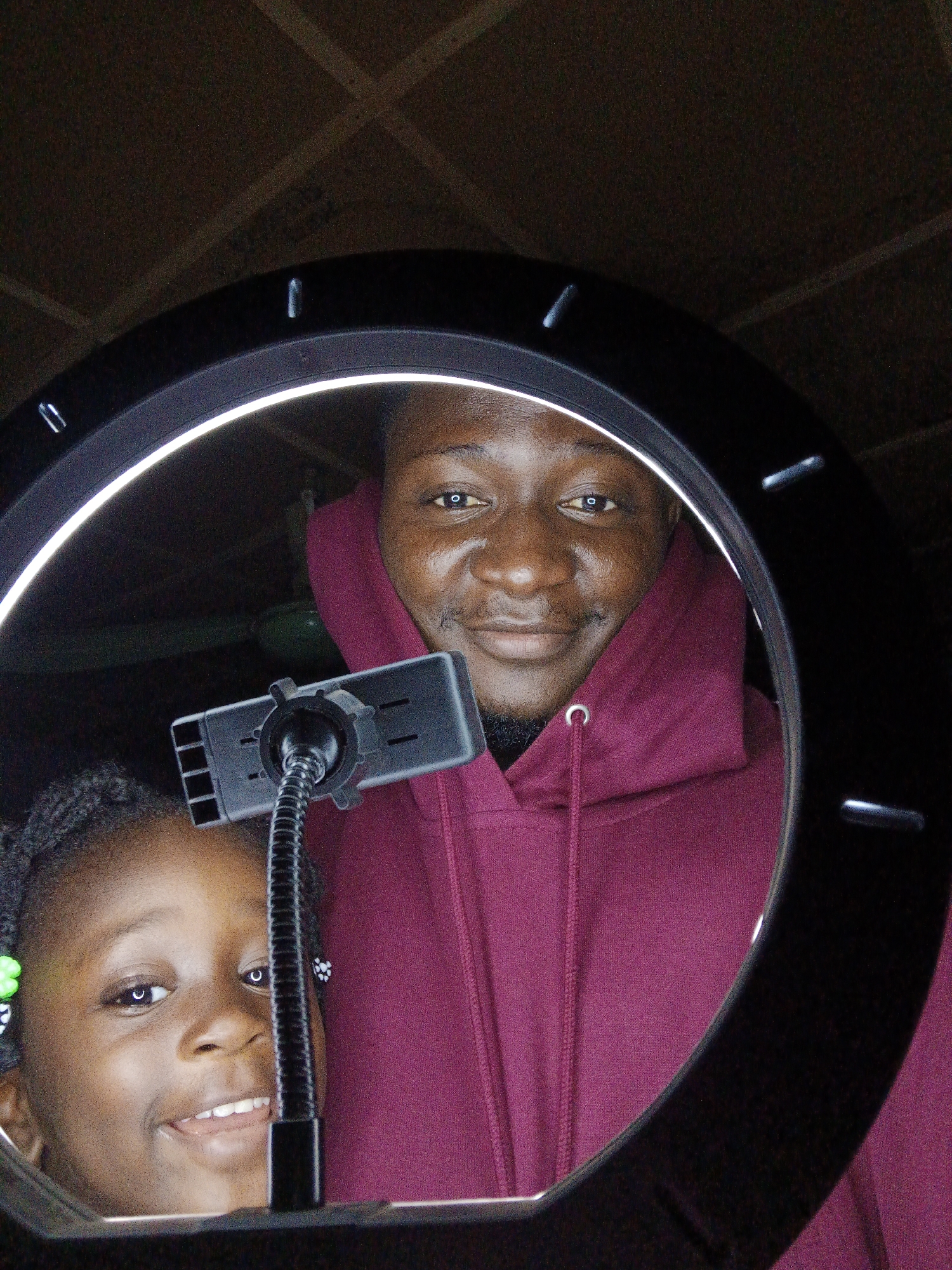A close-up, selfie-style photograph captures two individuals positioned within the illuminated circle of a ring light. The ring light itself is a prominent circular object, dark in color with a bright white interior glow. In the foreground and to the left, a young Black girl with braided hair is visible. She has dark eyes, a wide smile revealing her teeth, and is wearing small green and black flower-shaped earrings. In the center and to the right, a Black man with a short beard and mustache is positioned. He is wearing a dark reddish-purple hooded sweatshirt with white drawstrings. He is smiling faintly, with his eyes looking towards the camera. A phone mount with a flexible arm is situated between the two individuals, holding a phone that is not directly visible. The background appears to be a ceiling with a textured, dark brown surface, possibly wood paneling or a similar material. The overall lighting suggests an indoor setting, likely taken at night or in a dimly lit room, with the ring light providing the primary illumination. The environment is indistinct due to the focus on the subjects and the ring light.