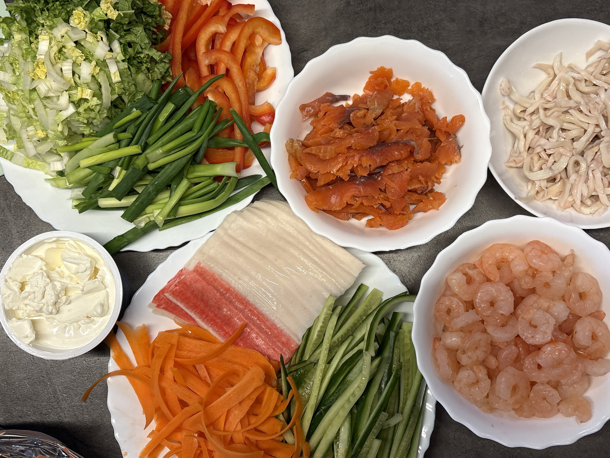 The image shows a top-down view of various food items arranged on a gray surface. The scene includes plates and bowls containing ingredients, possibly for making sushi or spring rolls.
Specifically, from left to right:
1. A white plate with shredded Chinese cabbage, cut scallions, and sliced red bell peppers.
2. A small white container filled with cream cheese.
3. A white plate holding julienned carrots, surimi sticks (crab sticks), and sliced cucumbers.
4. A white bowl containing cut, smoked salmon.
5. A white bowl filled with shredded, white meat, possibly chicken or squid.
6. A white bowl containing cooked shrimp.
The image provides no information about the location of the scene, and Birobidzhan, Russia, cannot be confirmed.