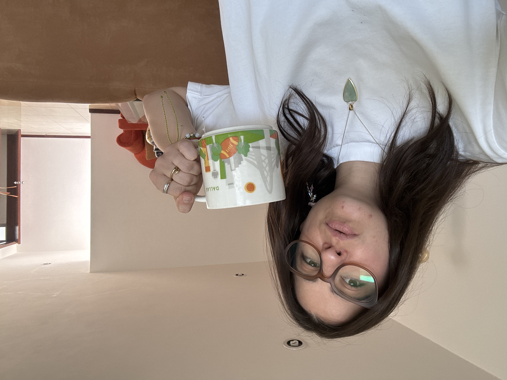 A woman with brown hair and glasses is holding a coffee mug. The mug has "DALLAS" printed on it along with city skyline images and a football. She is wearing a white t-shirt and jewelry, including a necklace with a green pendant. The background includes a beige wall, ceiling lights, and a partial view of a room with a brown couch. The image is rotated and inverted, making it appear as if the woman is standing on the ceiling. No evidence confirms that this is in Puebla, Mexico.