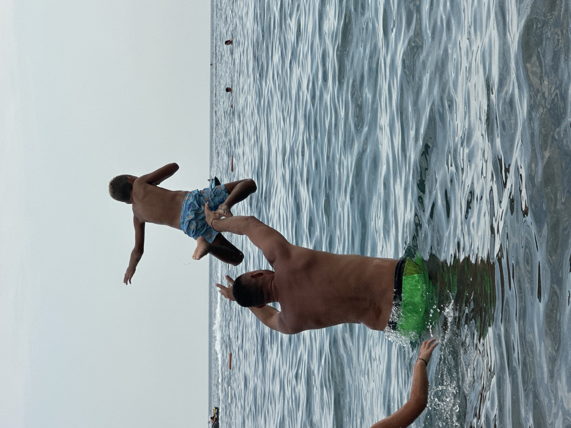 A father is throwing his son into the water at the beach in Boreti, Montenegro. The son is in mid-air, with his arms outstretched. The father is standing in the water, with his arms raised. There are other people in the water in the background, as well as a jet ski. The sky is overcast.
