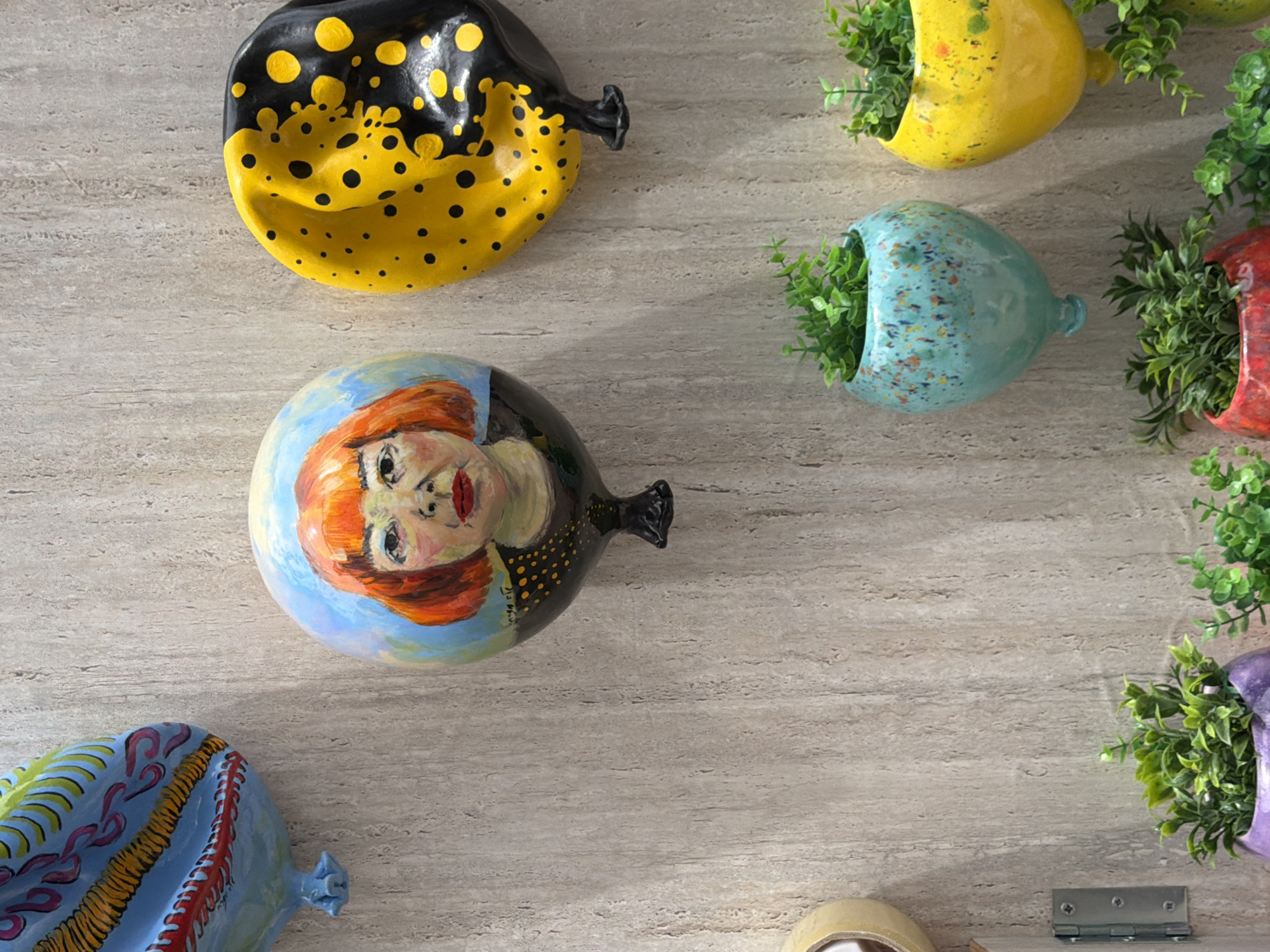 The image depicts a close-up view of a light gray, horizontally-striped surface adorned with multiple ceramic wall decorations. These decorations are shaped like deflated balloons and are painted with various designs and colors.
The top left balloon is black with yellow spots. Below it is a balloon painted with a portrait of a person with red hair. The bottom left shows a blue balloon with abstract patterns.
On the right side of the image are a variety of balloon-shaped planters. These planters are filled with green foliage. The planters are yellow, blue, and red. At the bottom right corner of the image, there is a partially visible purple planter with green foliage, and a small metal hinge on the wall. Also, visible at the bottom is a yellow tape roll.
There is no evidence to indicate that the image was taken in Tel-Aviv, Israel.