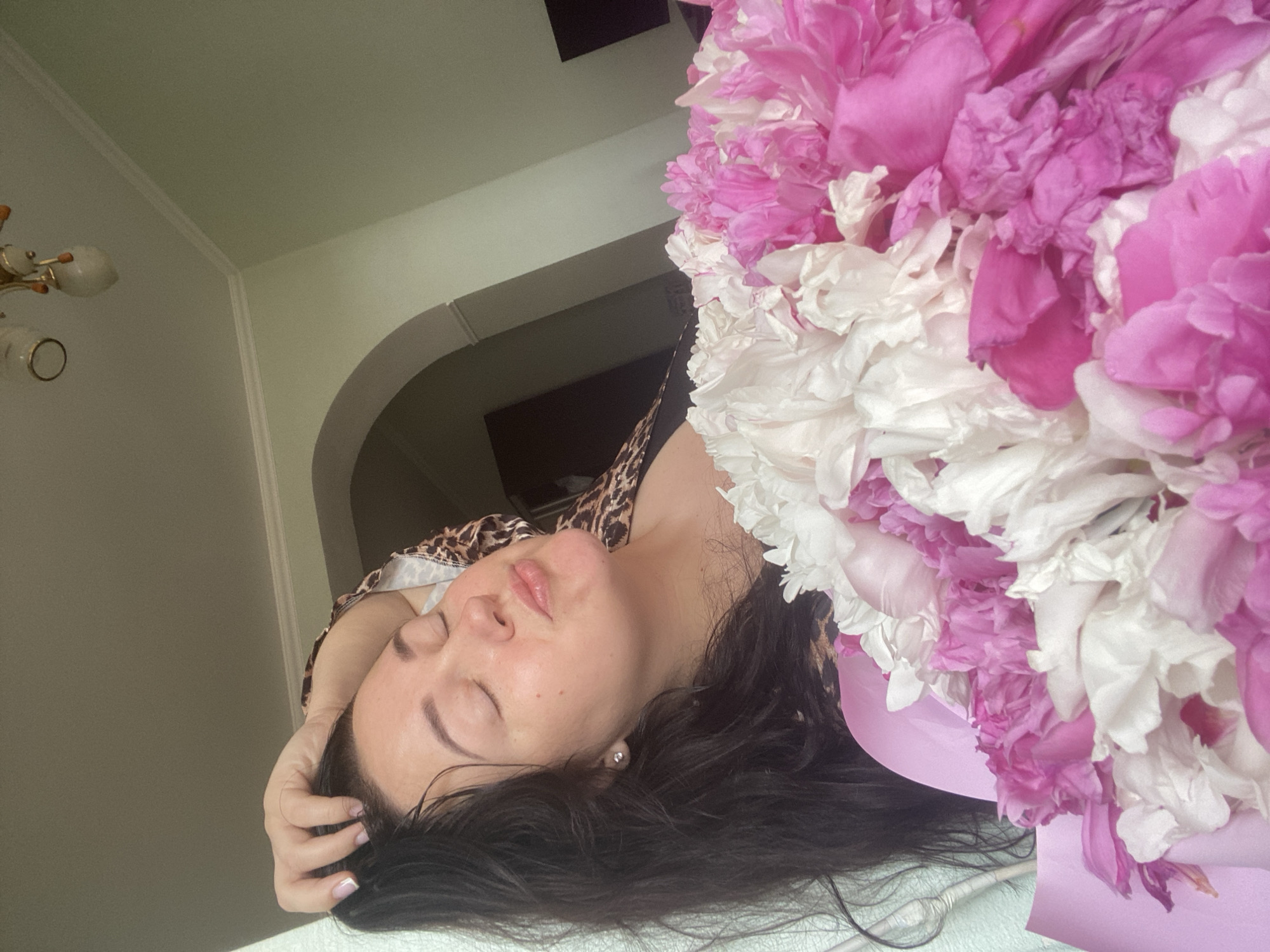 A woman with dark hair and fair skin is lying down next to a large bouquet of pink and white peonies. She is wearing a leopard print top. The background includes a wall with an archway and a light fixture. The location appears to be indoors.