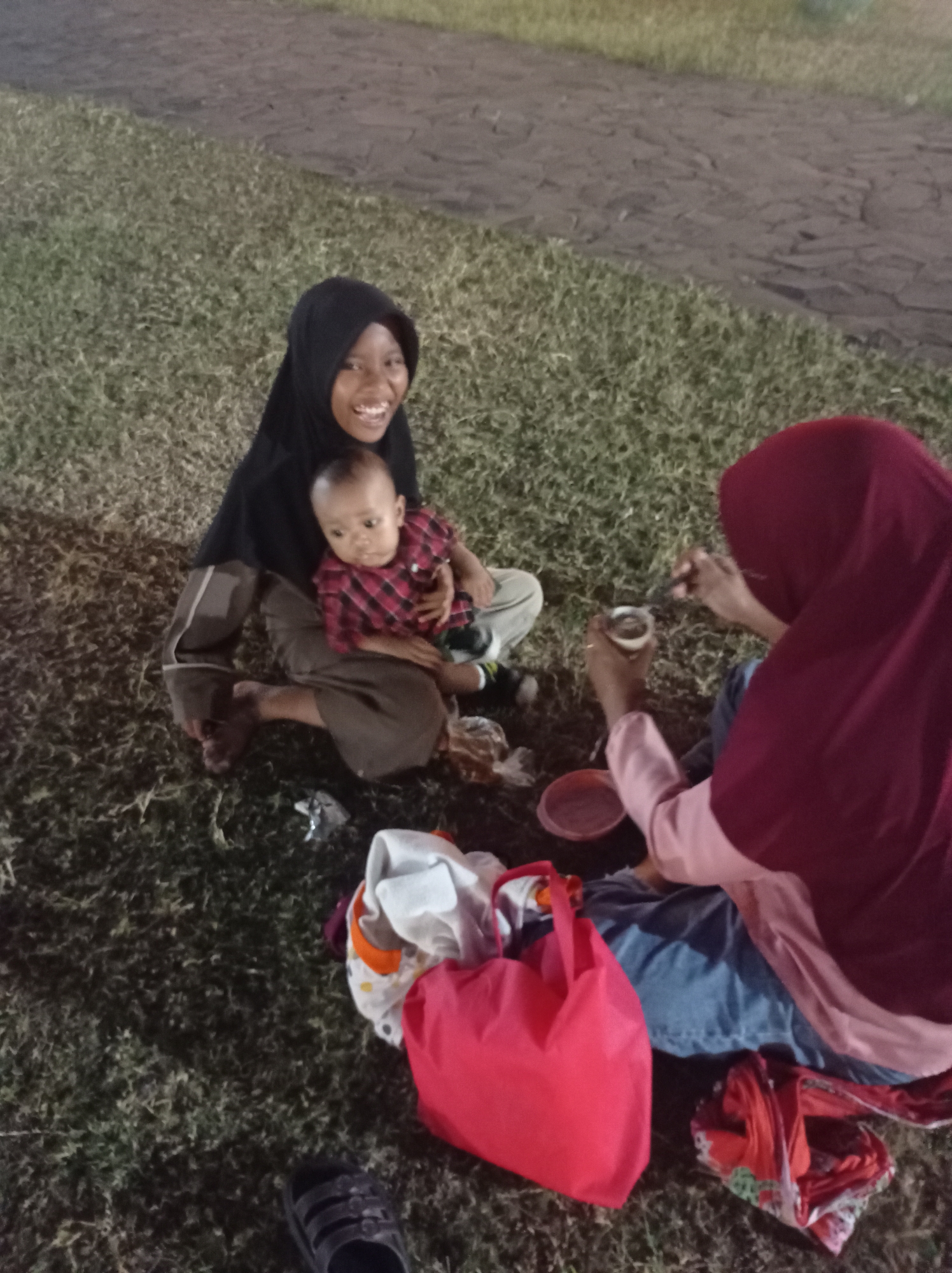 Two individuals, likely a woman and a child, are seated on a grassy area next to a paved pathway. The woman on the left, wearing a black hijab and a gray and white striped outfit, is holding a baby dressed in a red and black plaid shirt. She is smiling and looking towards the camera. The second individual, to the right and wearing a maroon hijab and a light pink top over blue jeans, is seated and appears to be eating from a small cup with a spoon. Several bags and items of clothing are scattered on the grass around them. The environment appears to be an outdoor public space, possibly a park, at dusk or in low light conditions, as indicated by the artificial lighting present in the background.