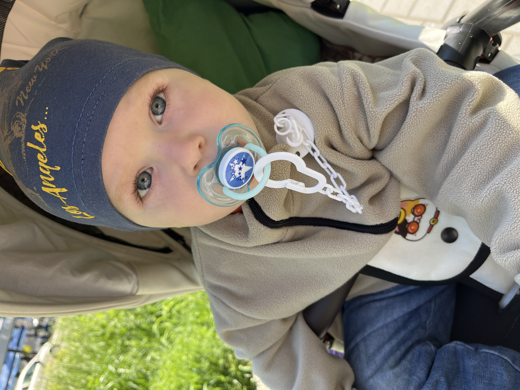 A baby, likely an infant or toddler, is pictured looking directly at the camera from a stroller or car seat. The baby has fair skin, bright blue eyes, and light eyelashes, framed by a dark blue beanie hat with yellow embroidered text that partially reads "New York" and "Los Angeles...". A blue and white pacifier, adorned with white stars, is in the baby's mouth and connected to a white pacifier clip with a chain.
The baby is dressed in a light beige, textured fleece jacket or blanket, and blue denim jeans are visible on the lower body. The setting appears to be outdoors during daytime, with soft, bright lighting. Out-of-focus green grass and some indistinct light-colored structures are visible in the background, suggesting a park or garden environment. The overall impression is one of calm and curiosity from the baby. While the location context is Chaikovsky, Russia, there are no specific visual cues within the image itself to confirm this. The baby's warm clothing (hat and fleece) suggests cooler weather, possibly spring or autumn.