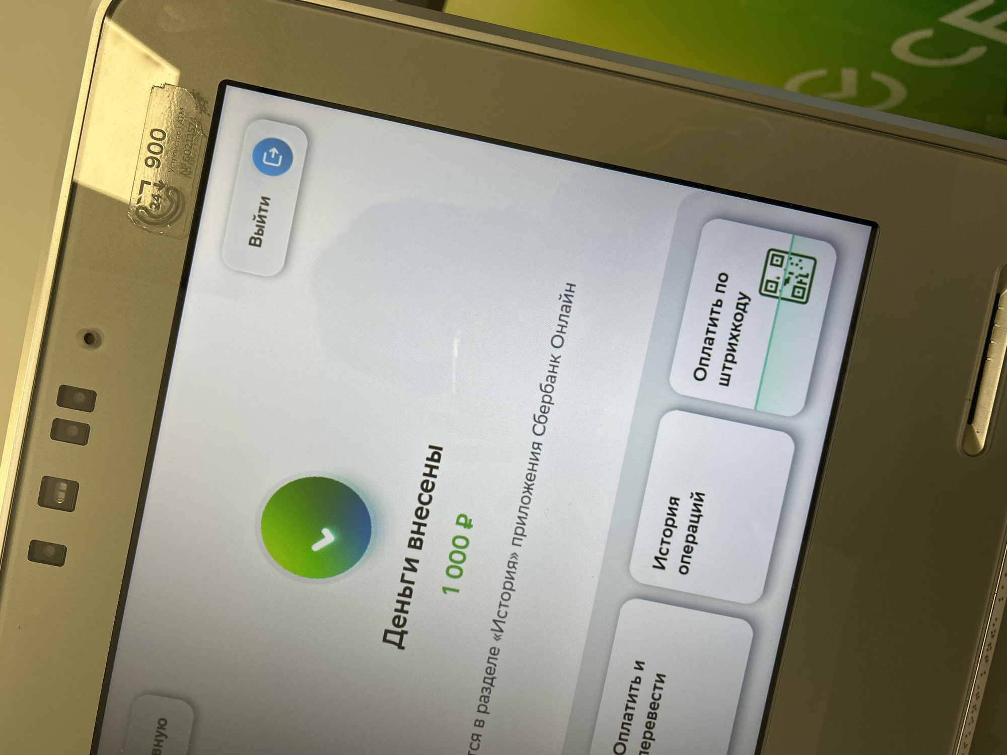 The image shows a close-up of a modern ATM screen displaying a confirmation message in Russian. The screen indicates "Деньги внесены" (Money deposited) and shows "1 000 ₽" (1000 rubles). Below this, it states that the transaction can be found in the "History" section of the Sberbank Online application.
The ATM itself appears to be a standard model, with several buttons or touch screen options visible. To the left, there's a button labeled "Оплатить и перевести" (Pay and transfer). In the center, there's a button for "История операций" (Transaction history). To the right, there's a button for "Оплатить по штрихкоду" (Pay by barcode), which also displays a QR code.
At the top of the ATM, there are several small indicators, possibly for sensors or cameras. A sticker with a phone number "900" and some other text is visible on the upper right side, suggesting it's a Sberbank ATM, likely in Russia. The background of the screen is predominantly white, with a green and blue circular graphic with a checkmark at the top. The overall environment appears to be indoors, possibly a bank branch or a public space equipped with an ATM. There are no people visible in the image. The metallic casing of the ATM is visible, reflecting some light. The image quality is good, allowing for clear reading of the text on the screen.
The specific location in Moscow, Russia, cannot be determined from the image alone.