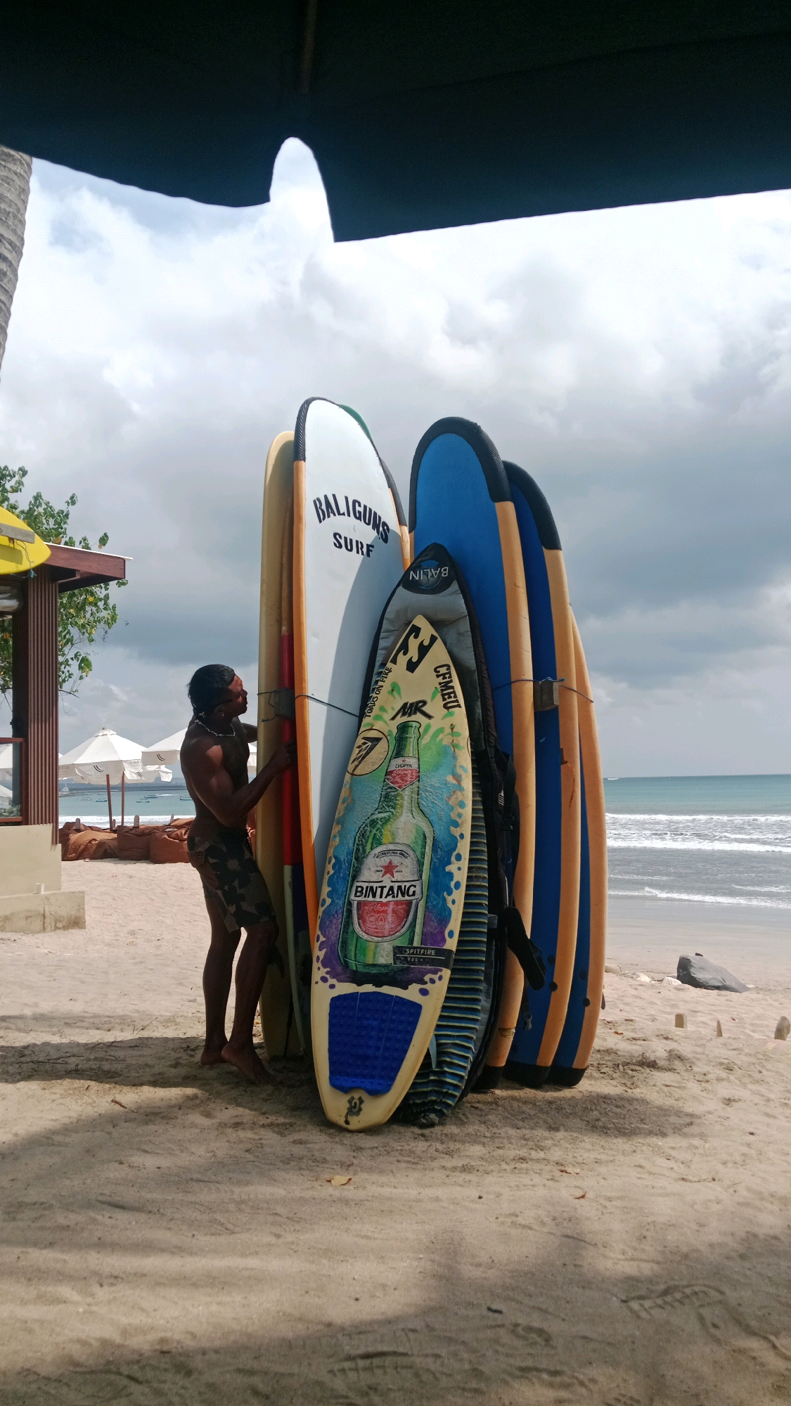 A shirtless man with dark hair and a tattoo on his chest stands on a sandy beach in Kuta, Indonesia. He is wearing camouflage shorts and is leaning against a rack of surfboards. The surfboards are propped upright and are of various colors and designs. One surfboard in the foreground features a colorful graphic of a Bintang beer bottle. In the background, the ocean is visible with gentle waves and a cloudy sky. There are also white umbrellas and a small building with a yellow awning visible on the beach. The scene appears to be in the late morning or early afternoon.