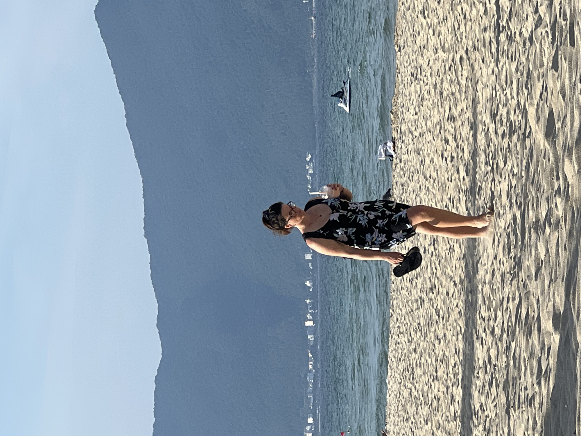 A woman walks on a sandy beach in Da Nang, Vietnam. She is wearing a black floral print dress and holding a drink in a clear plastic cup with a straw and a pair of black flip-flops. In the background, the ocean stretches towards a coastline dotted with buildings. Beyond the coastline, a large, forested mountain range rises against a clear blue sky. The water is a rippling blue, and a jet ski is visible in the distance. The sand is light brown and textured with footprints and shadows.