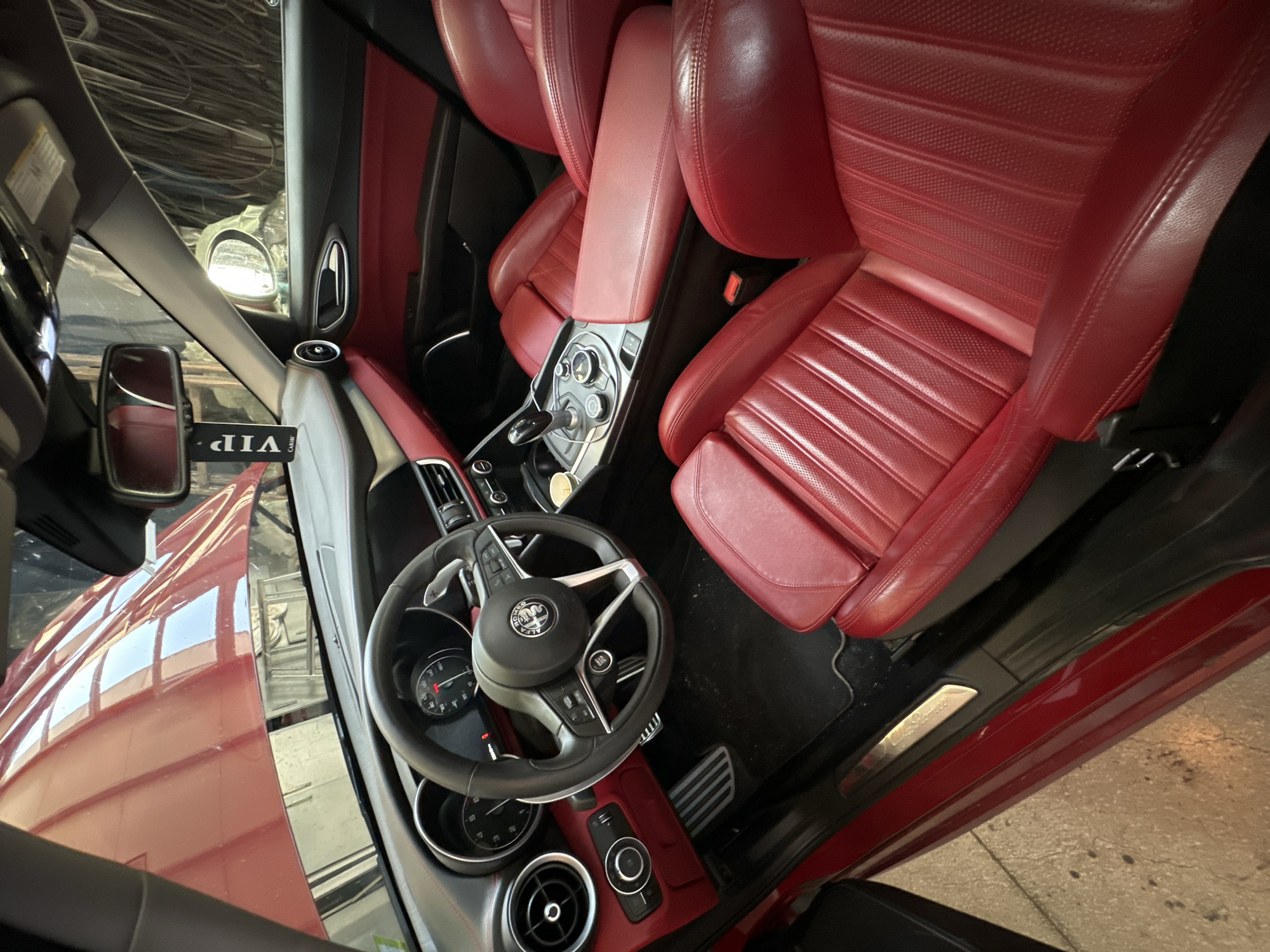 The image shows the interior of a red car, possibly an Alfa Romeo based on the badge on the steering wheel. The car is parked outdoors on a concrete surface, and the hood is open, revealing the engine bay. The interior features red leather seats, a black dashboard, and a steering wheel with paddle shifters. A "VIP" air freshener hangs from the rearview mirror. The overall scene suggests the car is either being detailed or is in a workshop. There are no people visible in the image. The location is identified as Unknown, Unknown.