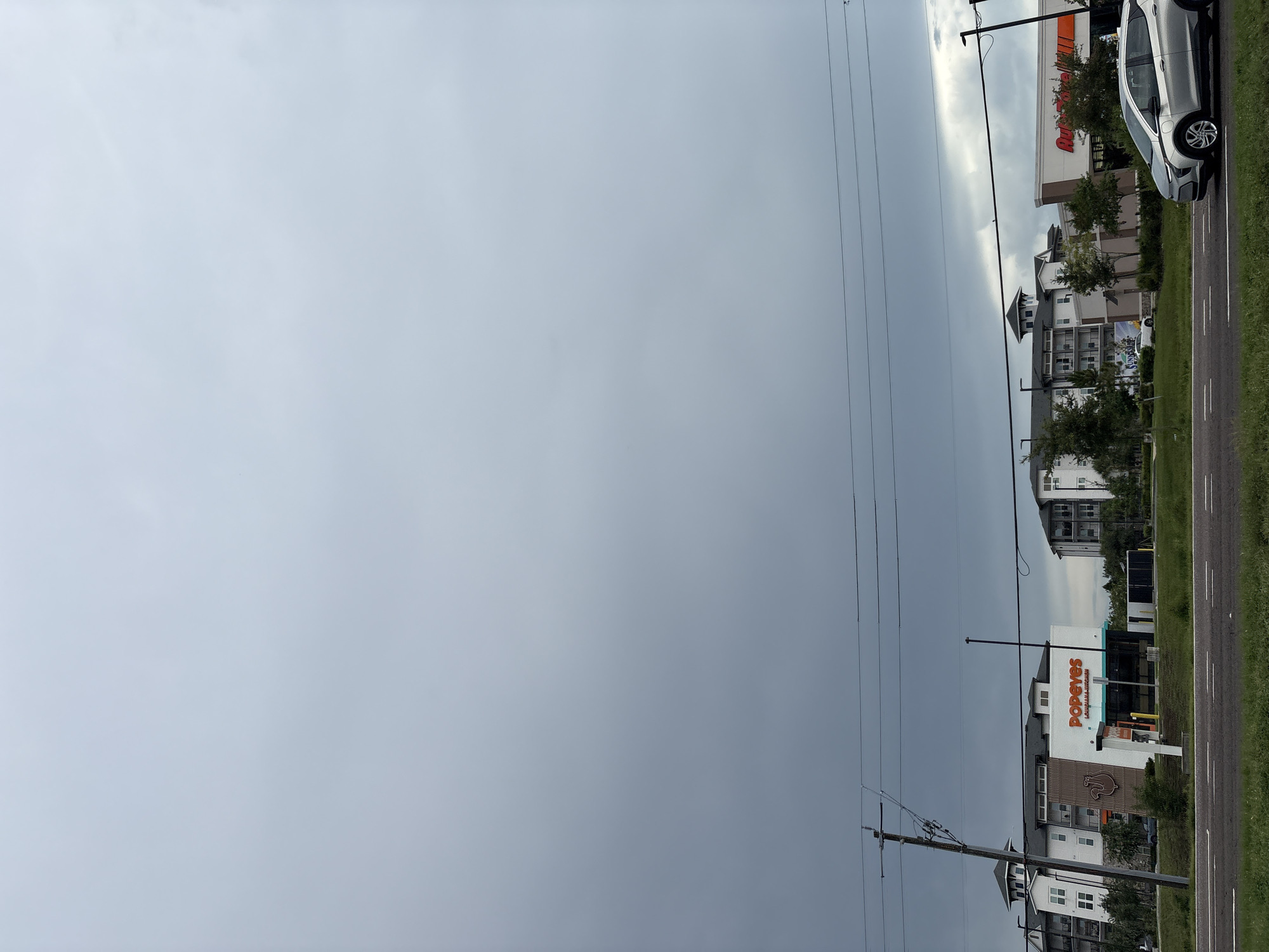 A silver electric car, a Chevrolet Bolt, is traveling on a road in front of a Popeyes Louisiana Kitchen and an AutoZone store. The buildings are located in Bradenton, Florida, United States. The sky is overcast, with dark clouds suggesting impending rain. The Popeyes restaurant has a large orange sign with the company logo, and the AutoZone store has a red and orange sign. There are several utility poles and power lines visible in the image. The road has white lane markings and a grassy shoulder.