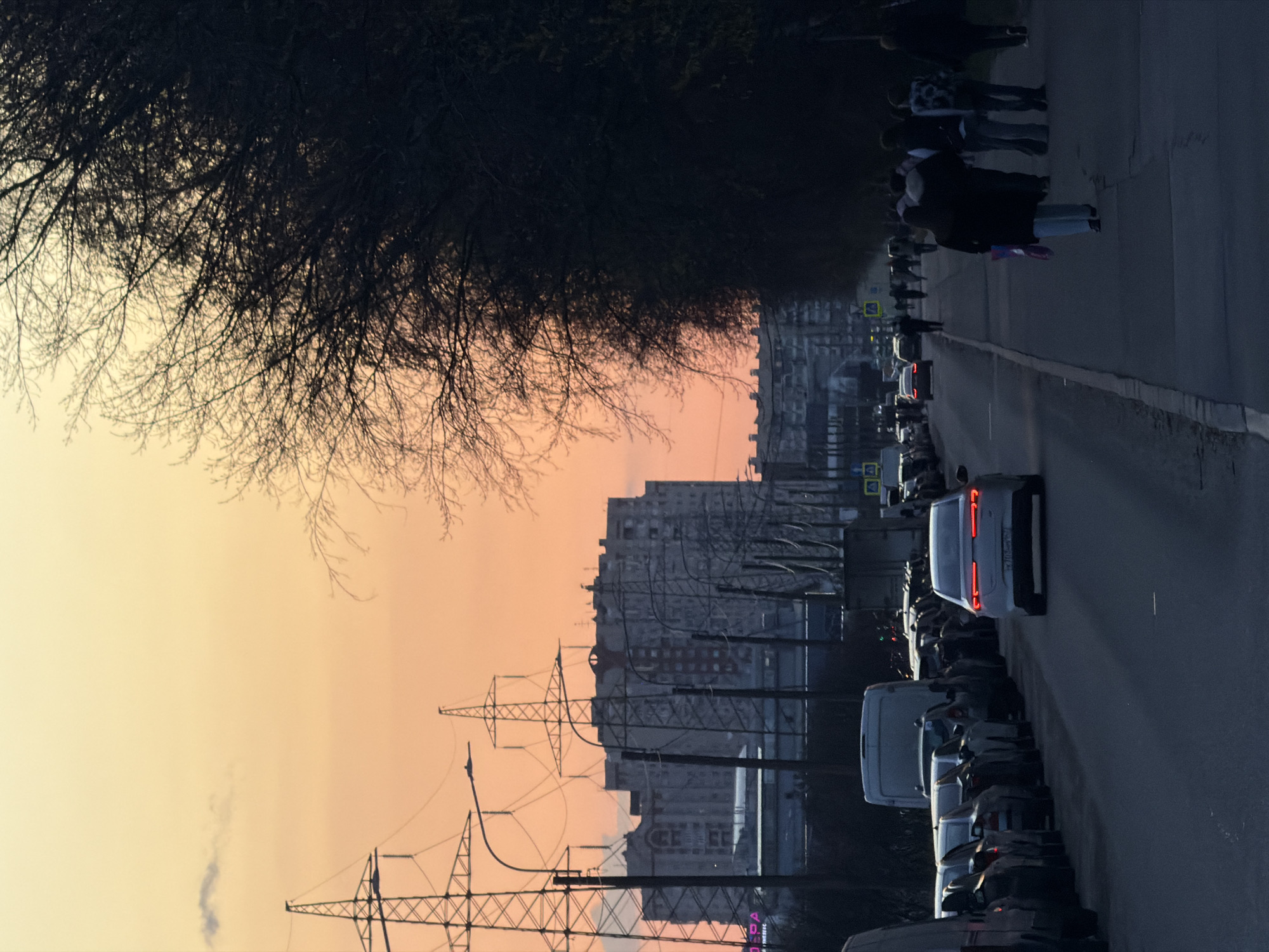 The image shows a street scene in Saint Petersburg, Russia, during what appears to be a late sunset or early twilight. The scene is viewed from a low angle, giving prominence to the sky, which is a gradient of orange and pale yellow. A bare tree with thin branches stretches across the upper left of the frame. Tall buildings are visible in the distance, partially obscured by power lines in the foreground.
A street runs alongside the buildings with parked cars on the right. A line of pedestrians, possibly heading home, can be seen walking on the sidewalk on the right. The street is lined with parked vans. The overall lighting suggests an urban environment transitioning from day to night.