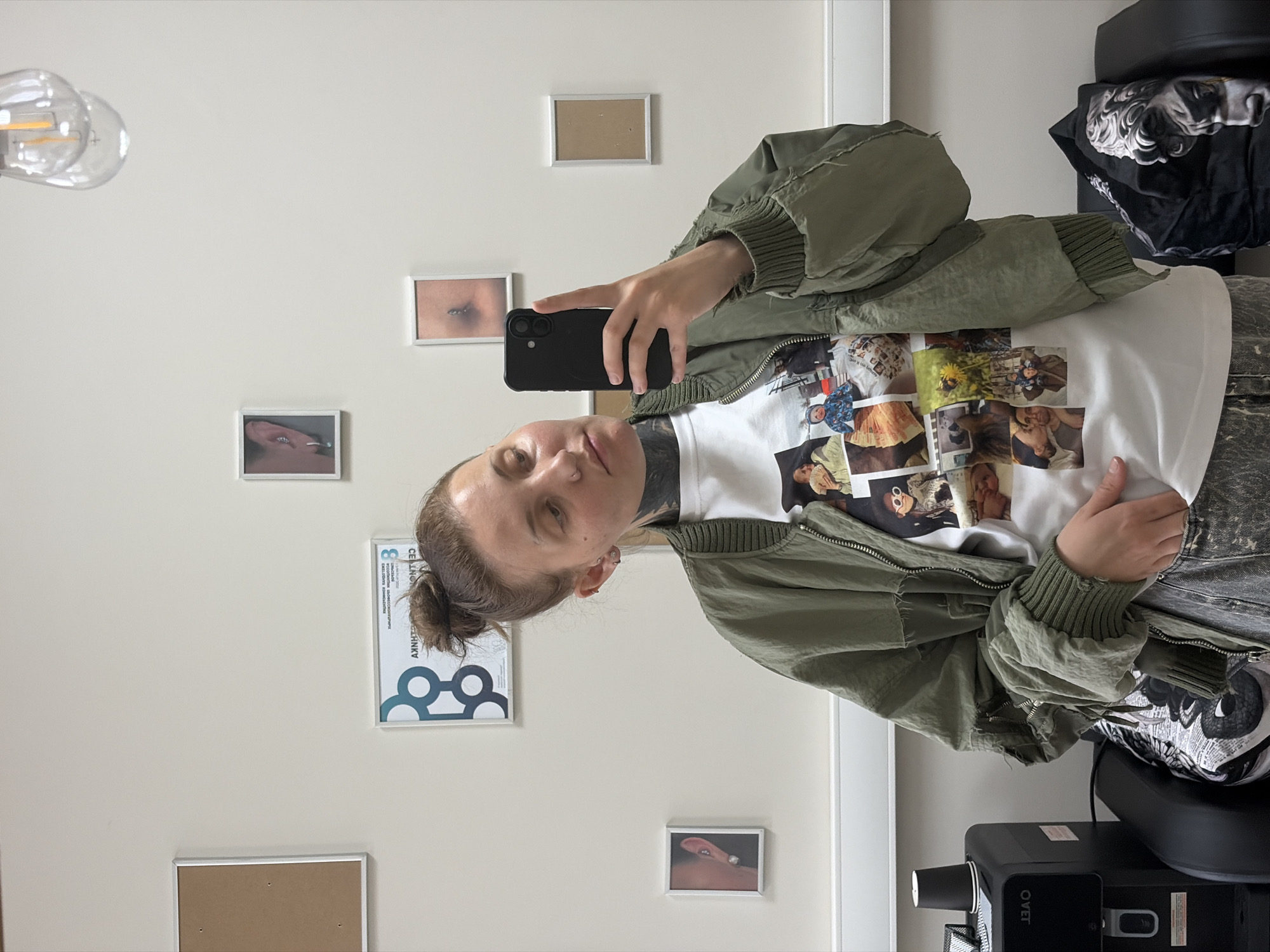 A vertical, full-body selfie is taken in a room with light-colored walls, possibly an art studio or gallery, given the framed pictures adorning the walls. The subjects are a young person with their hair tied up in a bun and visible tattoos on their neck. They are wearing a green bomber jacket over a white t-shirt featuring a collage of photographs. The person is holding a black smartphone to take the picture, with their right hand visible.
The background shows several small, framed pictures on the wall. Some appear to be abstract or artistic. One frame is empty. Another picture shows a close-up of an eye. A larger framed poster with geometric shapes and some text is also visible. To the right, part of a black chair and a black object with a white logo reading "OVET" are visible.
The lighting suggests daytime, with natural light possibly coming from the left. The overall mood is casual and personal, typical of a selfie. The room appears to be indoors, with clean, minimalist decor. There is no information about the weather.
Visible text includes "OVET" on the black object, and some text in Russian on the poster with geometric shapes. The text on the poster appears to be a title and some smaller explanatory text, but it is mostly illegible.