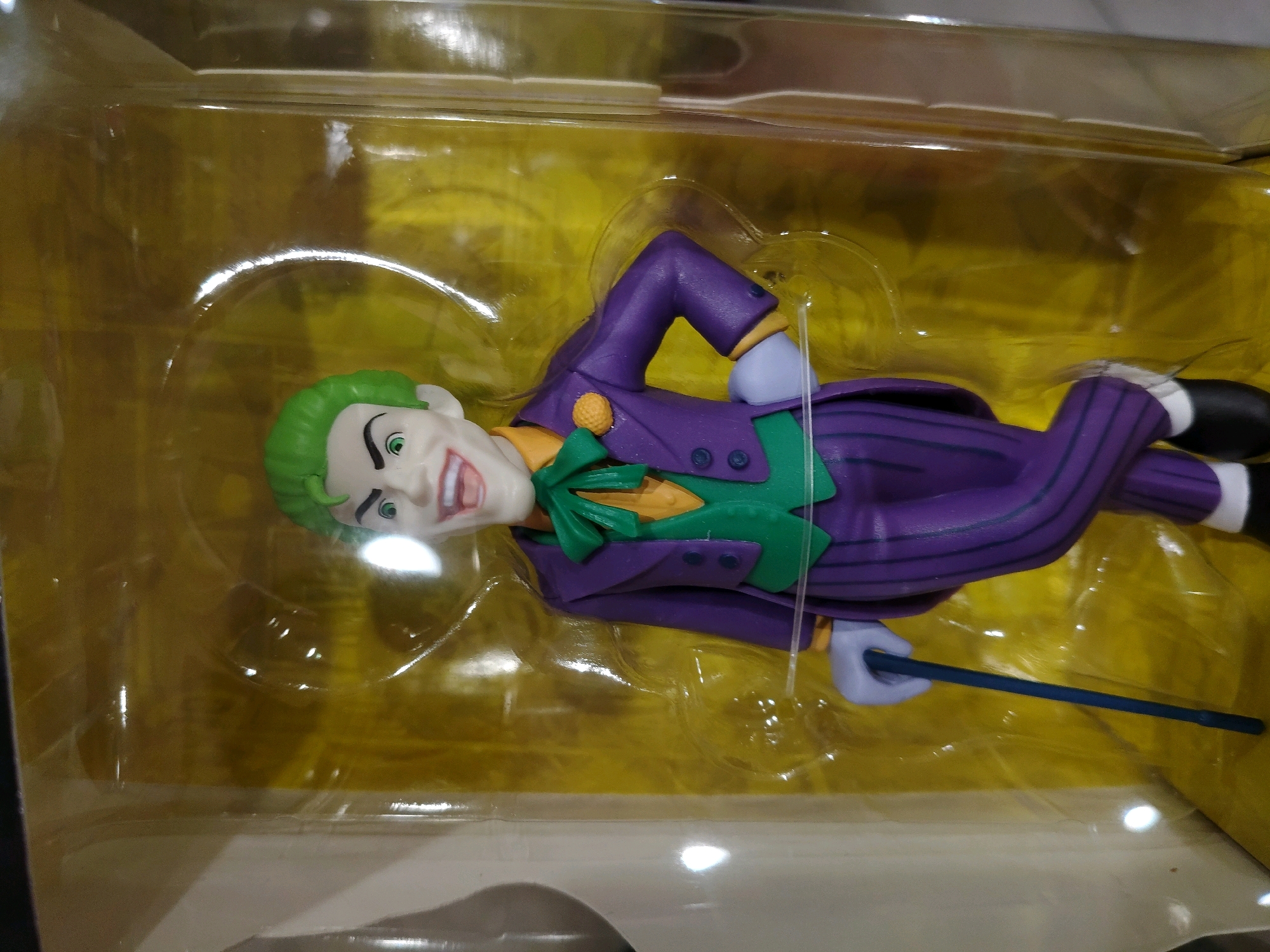 An action figure of the Joker, a Batman villain, lies horizontally in its plastic packaging. The figure is dressed in a purple suit with purple pinstripes, a green vest, and an orange shirt with a green bow tie. The Joker has green hair, a white face with exaggerated features, green eyes, and a wide, toothy grin. His left hand holds a thin, dark blue stick. The packaging has a yellow background. The image is taken from a slightly elevated angle, looking down at the figure.