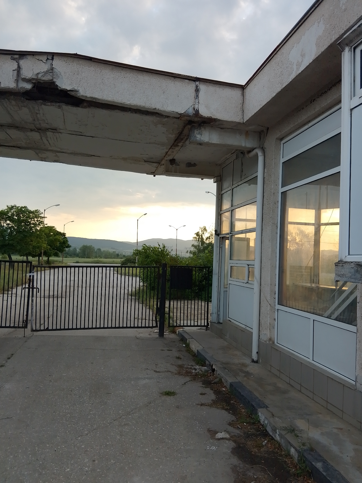 The image depicts an outdoor scene at what appears to be either sunrise or sunset, characterized by warm, low-angle light. In the foreground, a grey, worn concrete or asphalt surface extends, meeting a curb painted intermittently in black and grey, bordering a narrow strip of dirt and sparse vegetation.
To the right, a single-story structure, likely a former gatehouse or control booth, exhibits significant structural deterioration. Its exterior is a light-colored, possibly painted concrete or stucco, with multiple large, white-framed windows reflecting the golden light of the sky and distant trees. A white downspout runs vertically along the building's facade. The lower part of the wall is clad with grey tiles. The prominent concrete canopy or overhang above the entrance shows severe damage, including exposed rebar, large cracks, crumbling sections, and missing concrete soffit material. Rust stains are visible on the structural elements.
In the midground, a black metal gate, composed of vertical bars, is mostly closed, blocking a paved entranceway. The gate appears unlatched on its left side.
Beyond the gate, a paved road curves slightly into an open, somewhat overgrown landscape. Scattered green trees, including a large one on the far left, populate the area. Multiple streetlights with upward-curving arms are positioned along the road or in the field. In the distance, low hills or mountains are silhouetted against a sky featuring scattered clouds, which are illuminated by the warm, golden light of the sun nearing the horizon.
No individuals or active interactions are present in the scene. The overall impression is one of an aged or neglected facility in a semi-rural setting. The location is not identifiable beyond the visual elements presented.