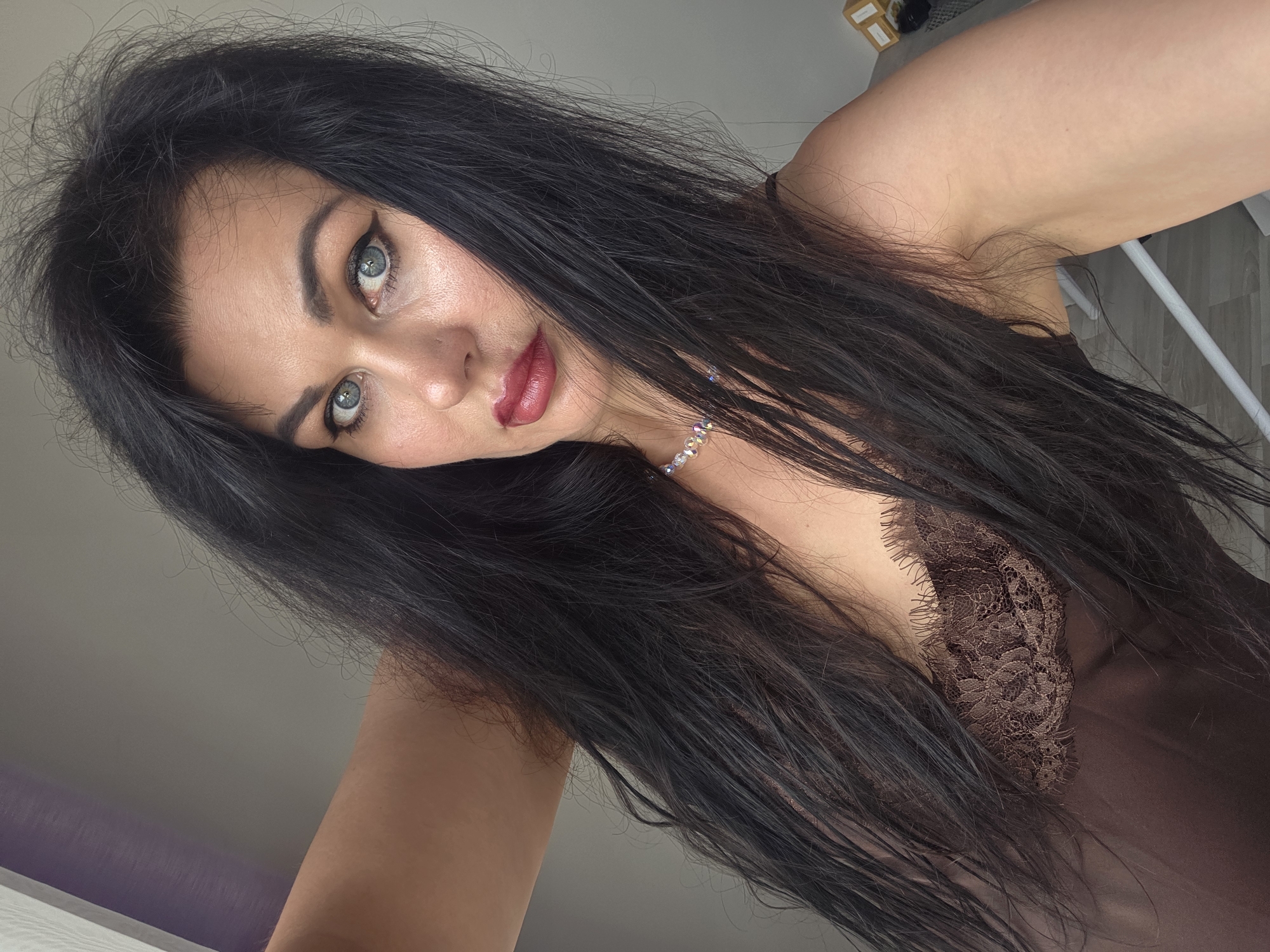 An adult female is captured in a selfie-style portrait, positioned indoors against a plain light gray wall. She has long, dark, voluminous hair that frames her face and cascades over her shoulders and chest. Her light blue eyes are accentuated with dark winged eyeliner, complemented by dark, defined eyebrows and glossy dark red lipstick. A choker-style necklace with clear, faceted beads is visible around her neck. She wears a brown or bronze-colored garment, likely a camisole or slip, featuring dark brown lace detailing with a floral pattern across the bust. Her raised right arm is partially visible in the upper left of the frame. In the background, on the far left, a narrow vertical strip of purple-colored surface is visible. On the far right, indistinct light-colored rectangular objects, possibly boxes, and a white linear object (a rail or rod) are partially visible. The specific location in the city of Sochi, Russia, cannot be determined from the visual information provided in the image.
