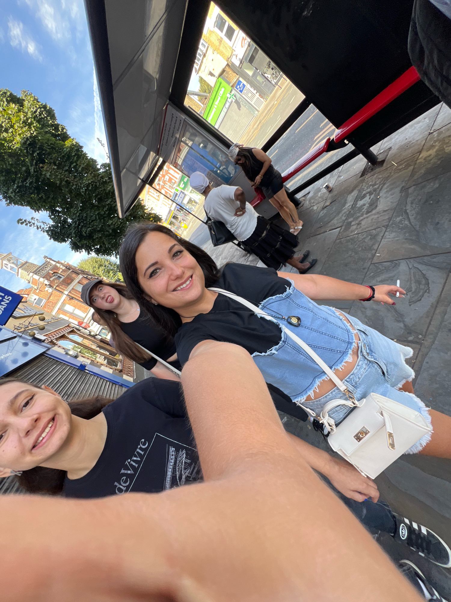 The image shows a group of people at a bus stop in London, UK. The main subjects are three young women, two of whom are in the foreground, smiling at the camera. The woman on the right is wearing a denim corset top and denim shorts, and is holding a cigarette. She has a white purse on her shoulder. The woman to her left is wearing a black t-shirt and a grey baseball cap, and is making a surprised face. In the background, other people can be seen waiting for the bus. The bus stop has a shelter with a red metal bench and a timetable display. The surrounding environment includes a street with traffic, buildings, and trees, suggesting an urban setting. The architectural style of the building with the clock tower visible in the background is brick, consistent with London's older buildings.