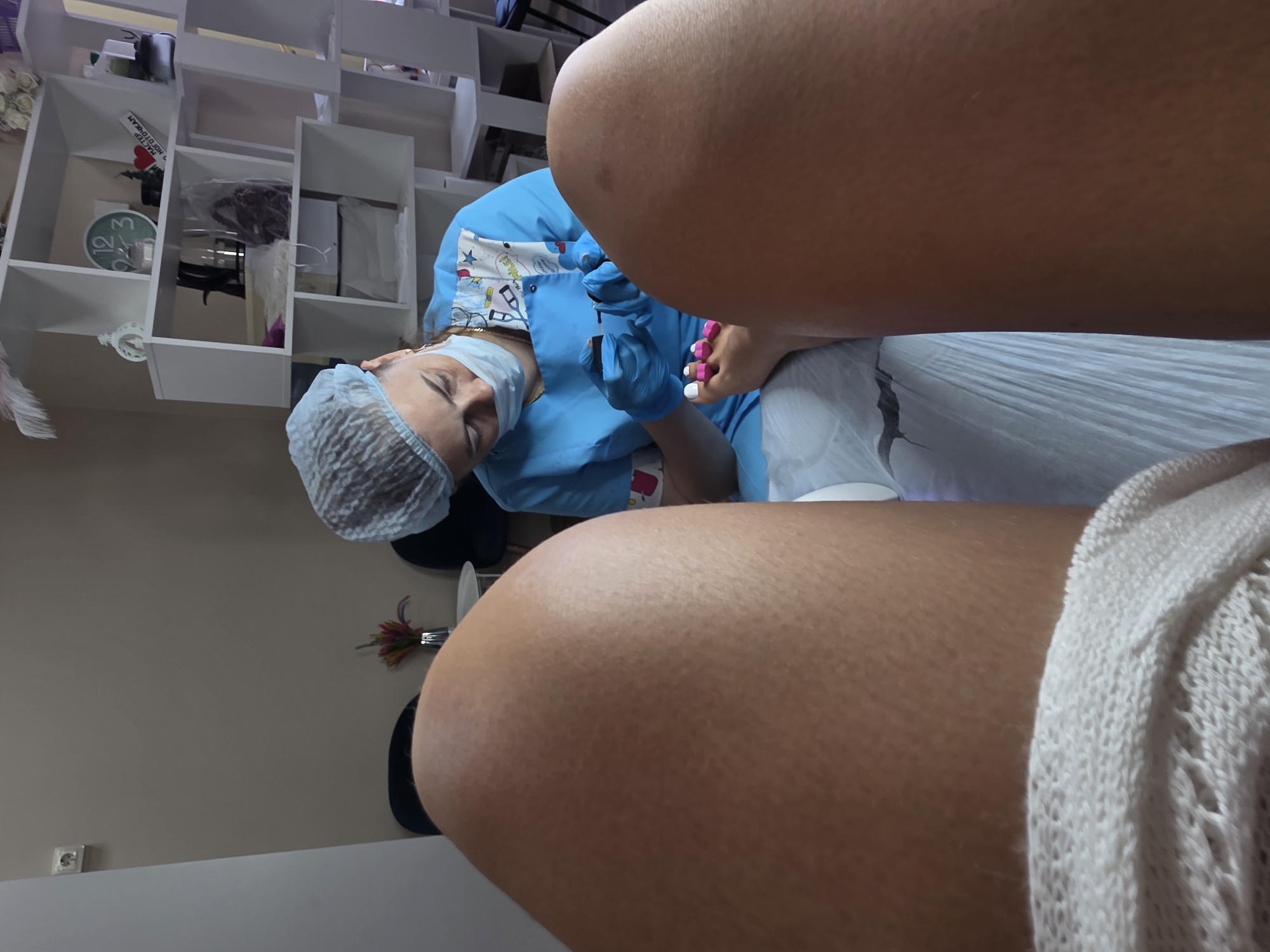 In an indoor setting, possibly a nail salon, a person is receiving a pedicure. The camera perspective is from the client's point of view, focusing on their legs and feet. A nail technician, wearing a blue uniform, hairnet, and mask, is meticulously painting the client's toenails white. The client's feet are positioned with pink toe separators. In the background, shelves are visible, containing various items, including a decorative clock. The lighting is neutral, and the overall atmosphere suggests a professional service. The location is stated to be Sochi, Russia.