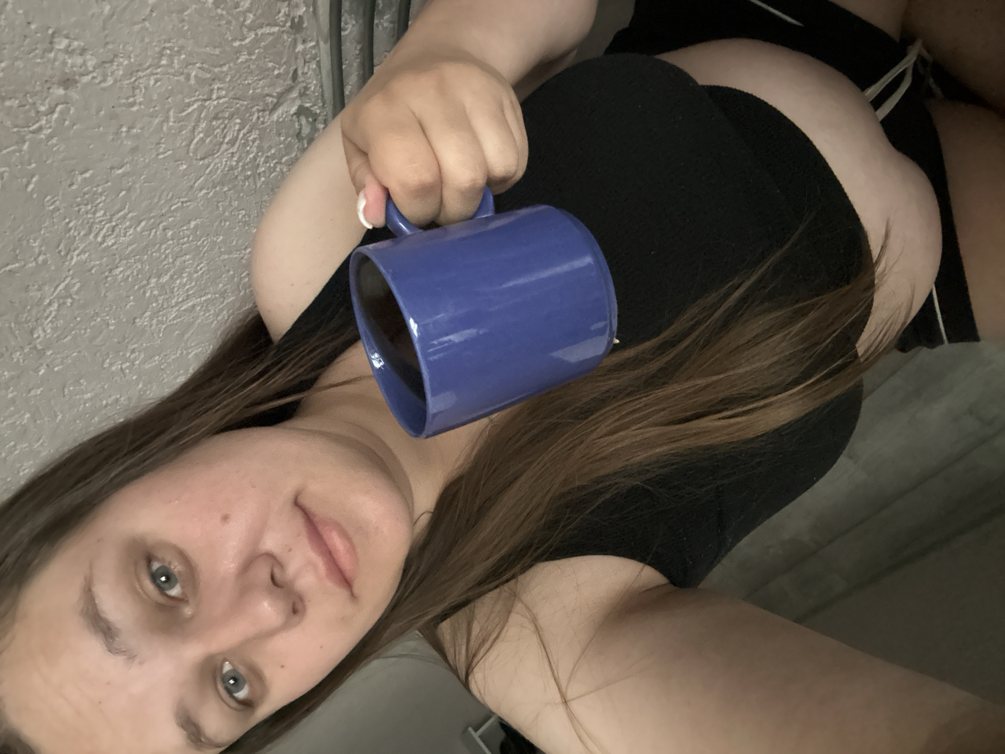 A young adult female is depicted in a medium-shot selfie, positioned in the city of Chaikovsky, Russia. She has long, straight, brown hair parted slightly to her left, reaching her chest. Her face is light-toned with light blue eyes, prominent under-eye circles, a small mole on her right cheekbone, and full, light pink lips. She maintains a neutral to slightly soft expression while looking directly at the camera. She is wearing a black sleeveless crop top that exposes her midriff, and black shorts with white stripes along the outer seams. Her left hand, featuring a white French tip manicure, holds a solid blue ceramic mug containing a dark liquid, presumably coffee or tea. The background consists of a light grey wall with a distinct, rough stucco-like texture. Lighting is even, suggesting an indoor setting.