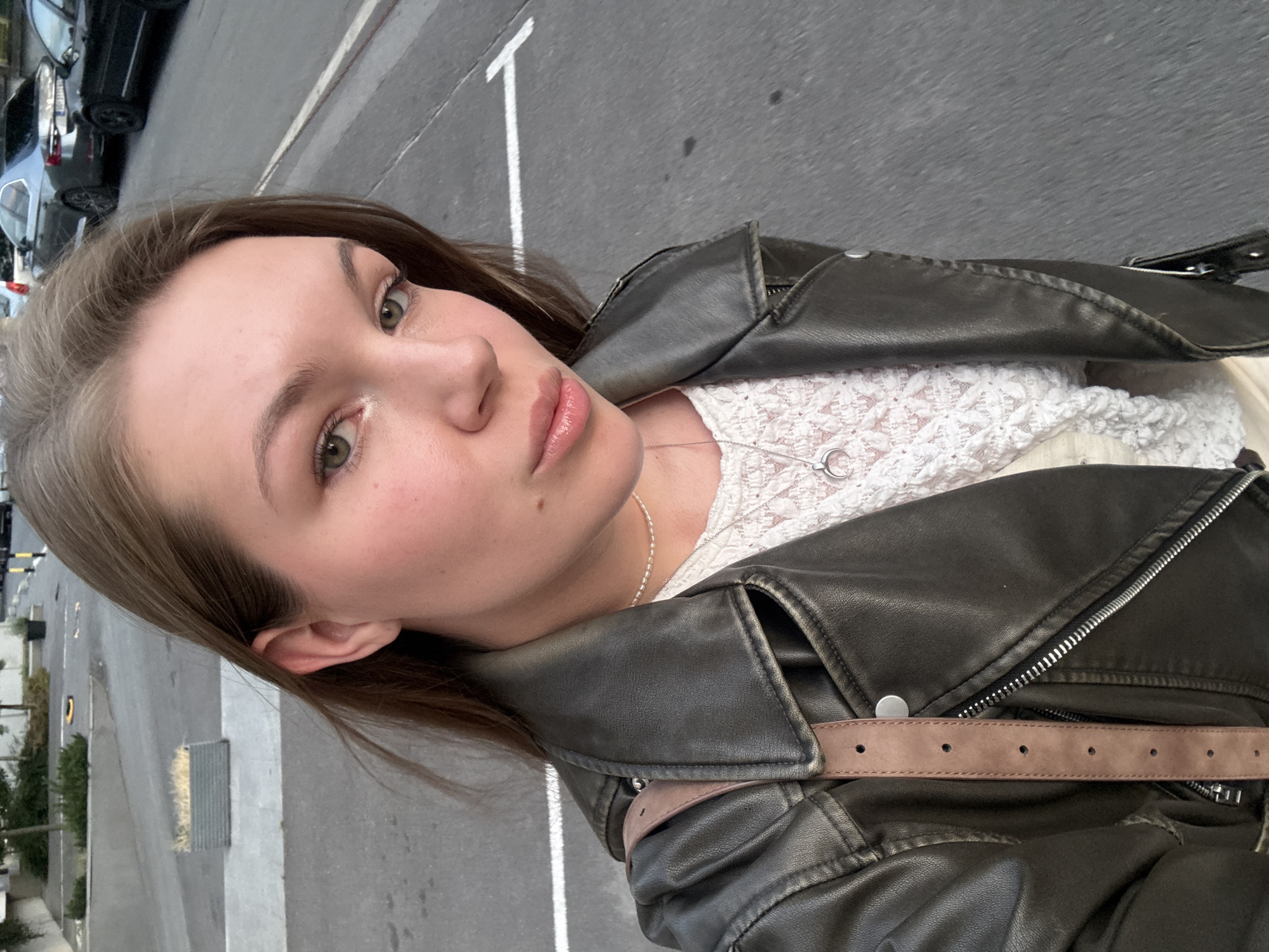 A young woman with fair skin, brown hair, and green eyes takes a selfie outside. She is wearing a white lace top and a black leather jacket with a brown strap. The background shows a city street with cars parked along the side and a white parking line. The image appears to be taken in Vozdovac Urban Municipality, Serbia.