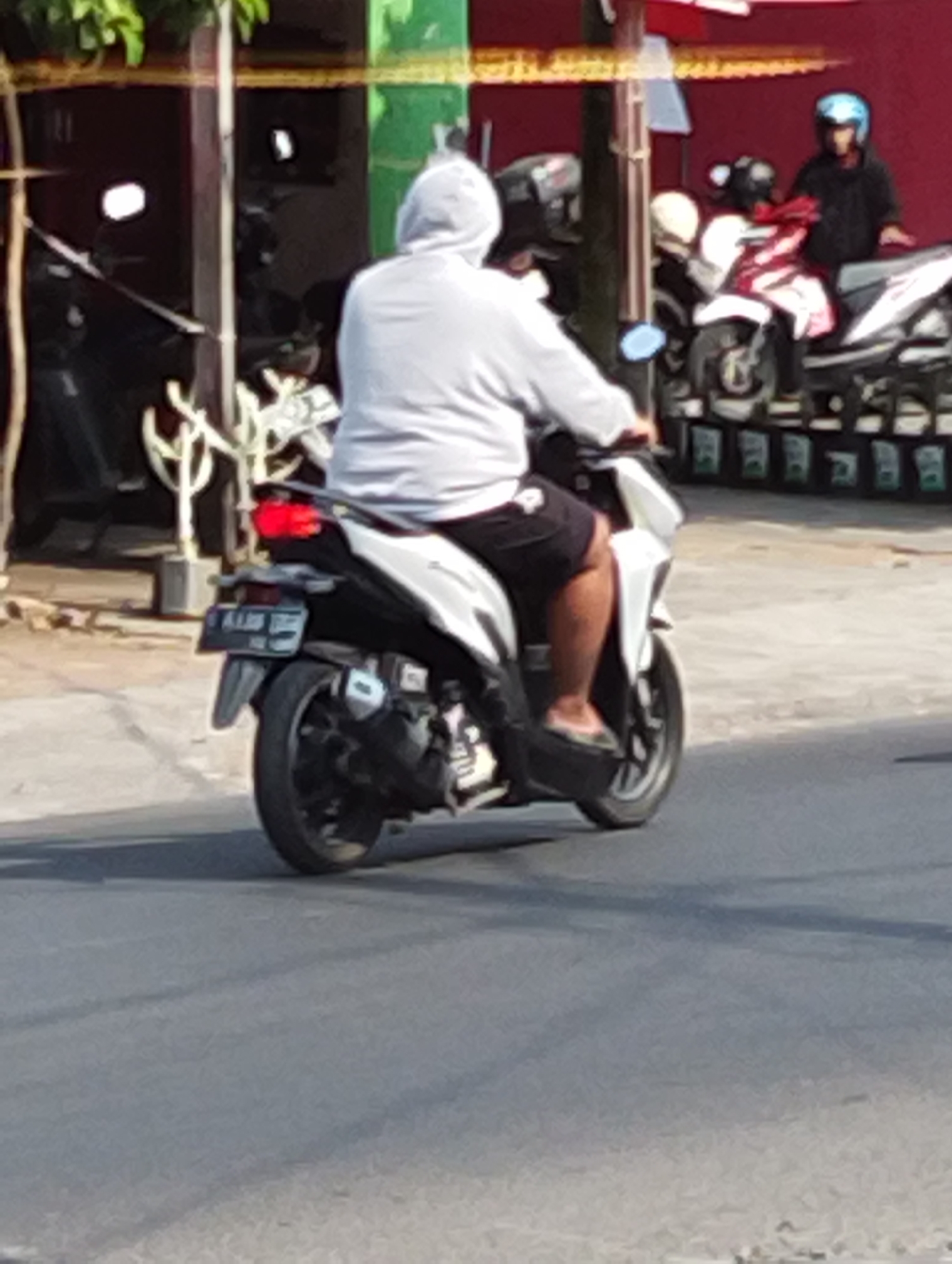 In Manaruwi, Indonesia, a person wearing a light gray hooded sweatshirt and black shorts is riding a white scooter. The scooter has a black license plate with white text. In the background, another scooter is visible with two people on it, one wearing a blue helmet. The scene appears to be on a street with buildings and trees in the background. The lighting suggests it is daytime.