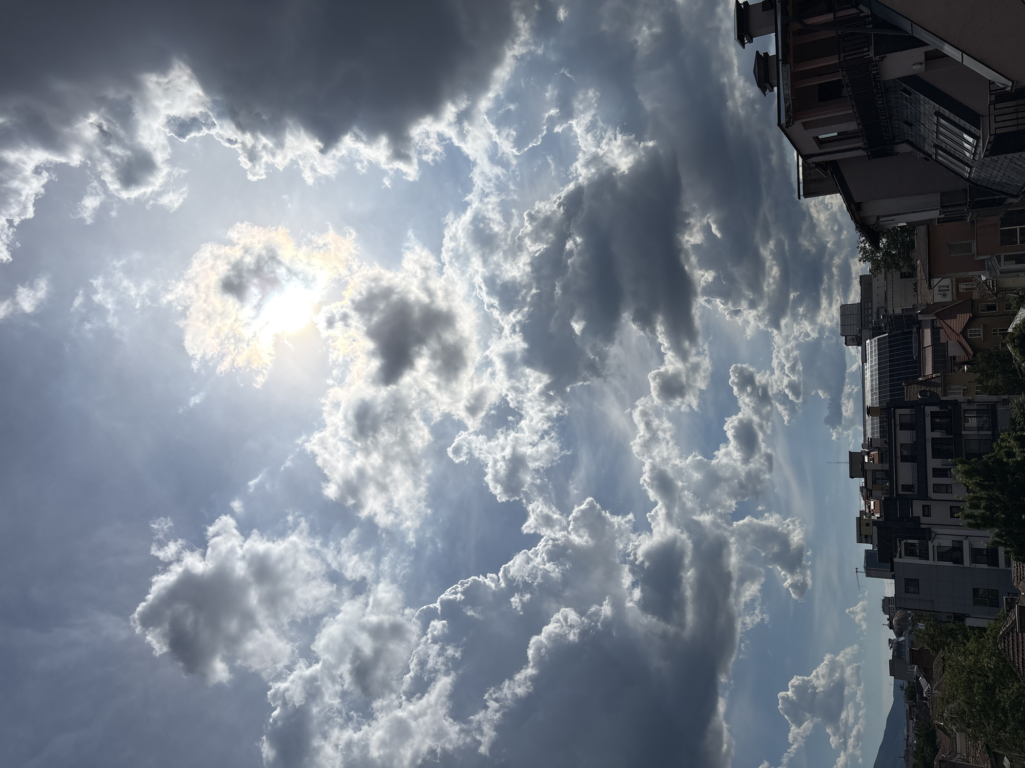 A wide-angle, vertical photograph captures a dramatic sky filled with a mix of bright, illuminated cumulus clouds and darker, more ominous storm clouds. The sun is partially obscured by clouds in the upper right quadrant, creating a bright halo effect and casting rays of light. Below the sky, the rooftops and upper stories of buildings in a city are visible, suggesting an urban environment. The architecture appears to be a mix of older tiled roofs and modern apartment buildings with balconies. The scene is illuminated by natural sunlight, indicating daytime. The location is identified as Sofia, Bulgaria.