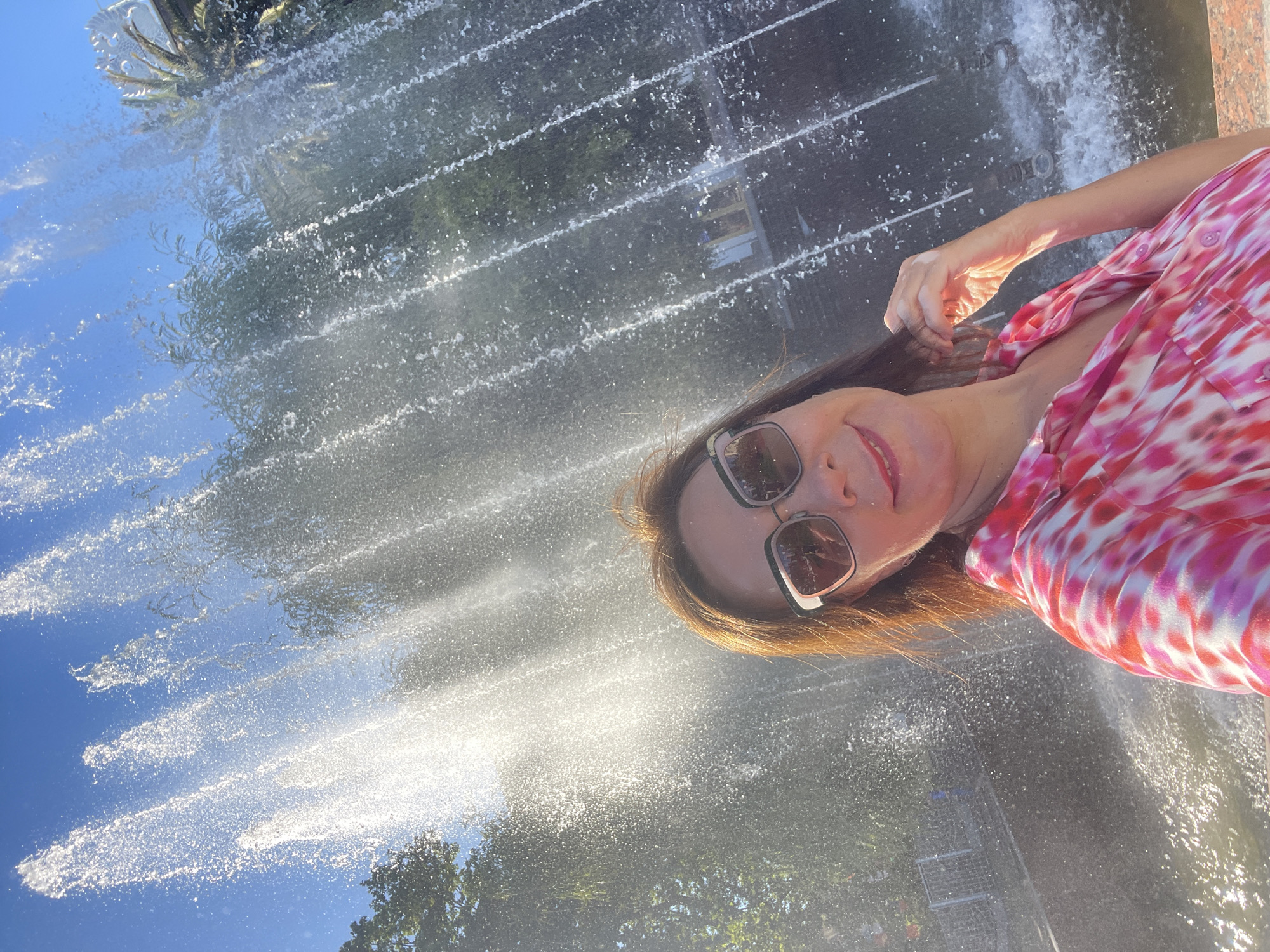 The image shows a woman taking a selfie in front of a fountain in Sochi, Russia. The woman is wearing sunglasses and a pink and white tie-dye shirt. She is smiling and appears to be enjoying herself. The fountain is spraying water in a curtain-like fashion, and the sun is shining brightly, creating a bright, airy atmosphere. The background shows trees and a clear blue sky. The time of day appears to be daytime, likely a sunny afternoon, given the bright lighting and shadows. The overall impression is one of a joyful and refreshing moment captured during a visit to this Russian city.