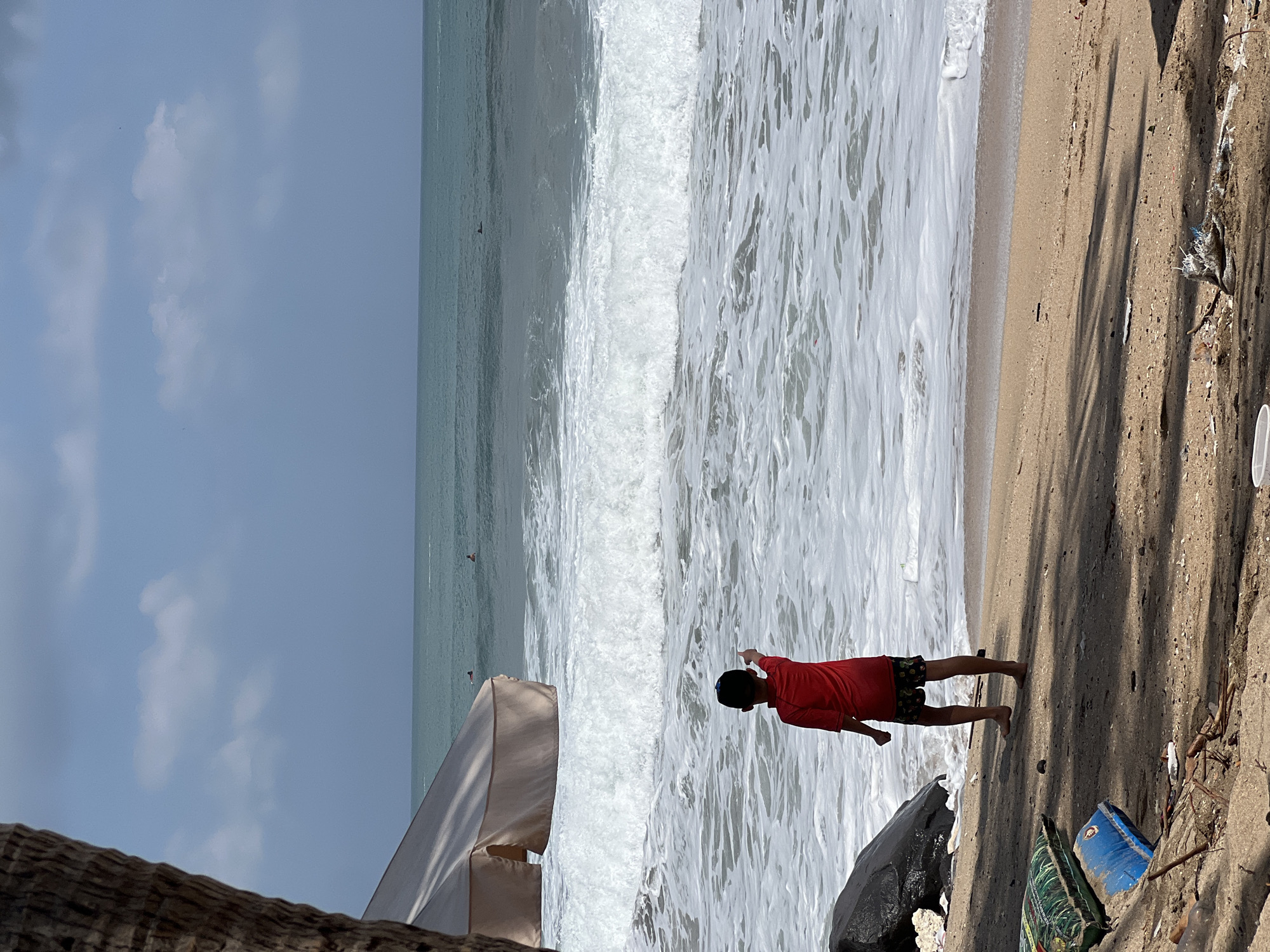 A young boy in a red shirt and dark shorts stands on the sandy shore of Kuta Beach, Indonesia, facing the ocean and pointing with his right hand. Large waves are breaking on the beach, with white foam washing ashore. In the distance, several surfers can be seen in the water. To the left of the boy, a large dark rock sits on the sand, and behind it, part of a beach umbrella is visible. The beach is littered with various pieces of debris, including plastic bags and other trash. The sky above is partly cloudy.