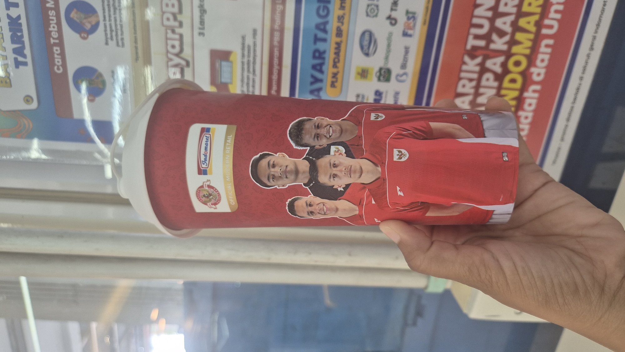 The image shows a person holding a red and white disposable cup with a picture of four Indonesian soccer players in red jerseys. The cup also features the Indomaret logo and the text "OFFICIAL MODERN RETAIL". The background appears to be a blur of vertical lines, possibly indicating a window or a display with multiple panes. To the right of the cup, a poster is partially visible with text in Indonesian, including "BAYAR PBB" and "AYAR TAGIHAN". The lighting suggests an indoor environment, possibly a store or a cafe, with light coming from the left. The overall impression is that the photo was taken inside a commercial establishment in South Jakarta, Indonesia, likely an Indomaret convenience store, given the branding on the cup.