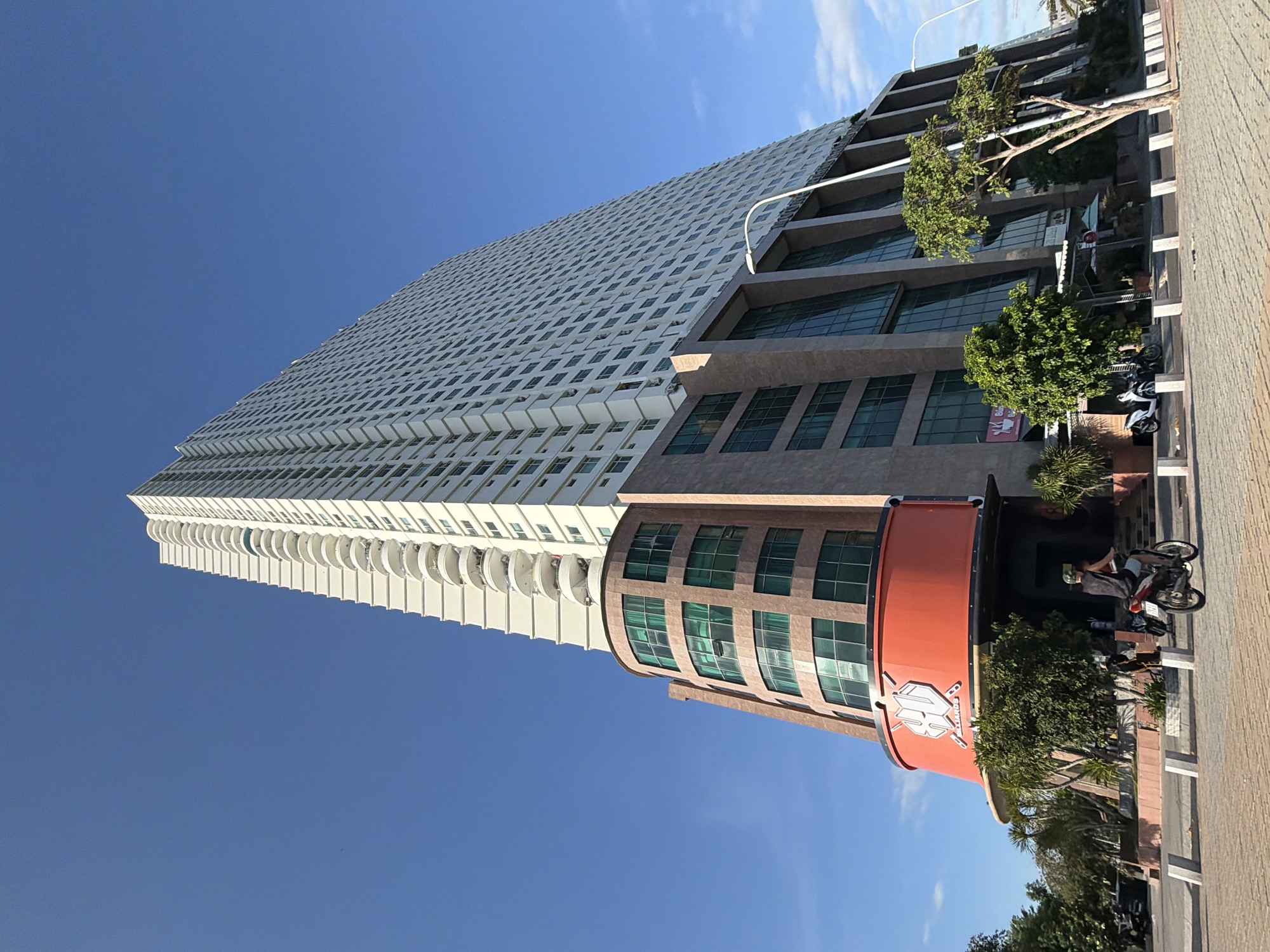 This image captures a low-angle view of the Havana Hotel in Nha Trang, Vietnam, a prominent skyscraper located on Tran Phu Street along the city's coastline. The hotel features a distinctive white, tiered structure with numerous balconies, contrasted by a modern, curved base with green-tinted windows. The building stands against a clear, bright blue sky. In the foreground, a paved walkway is visible with a motorcyclist in motion. There are also parked motorcycles and lush green trees surrounding the building's base, indicating a tropical urban setting. The building's lower section has a prominent orange circular structure with a logo that appears to read "HD BILLIARDS."