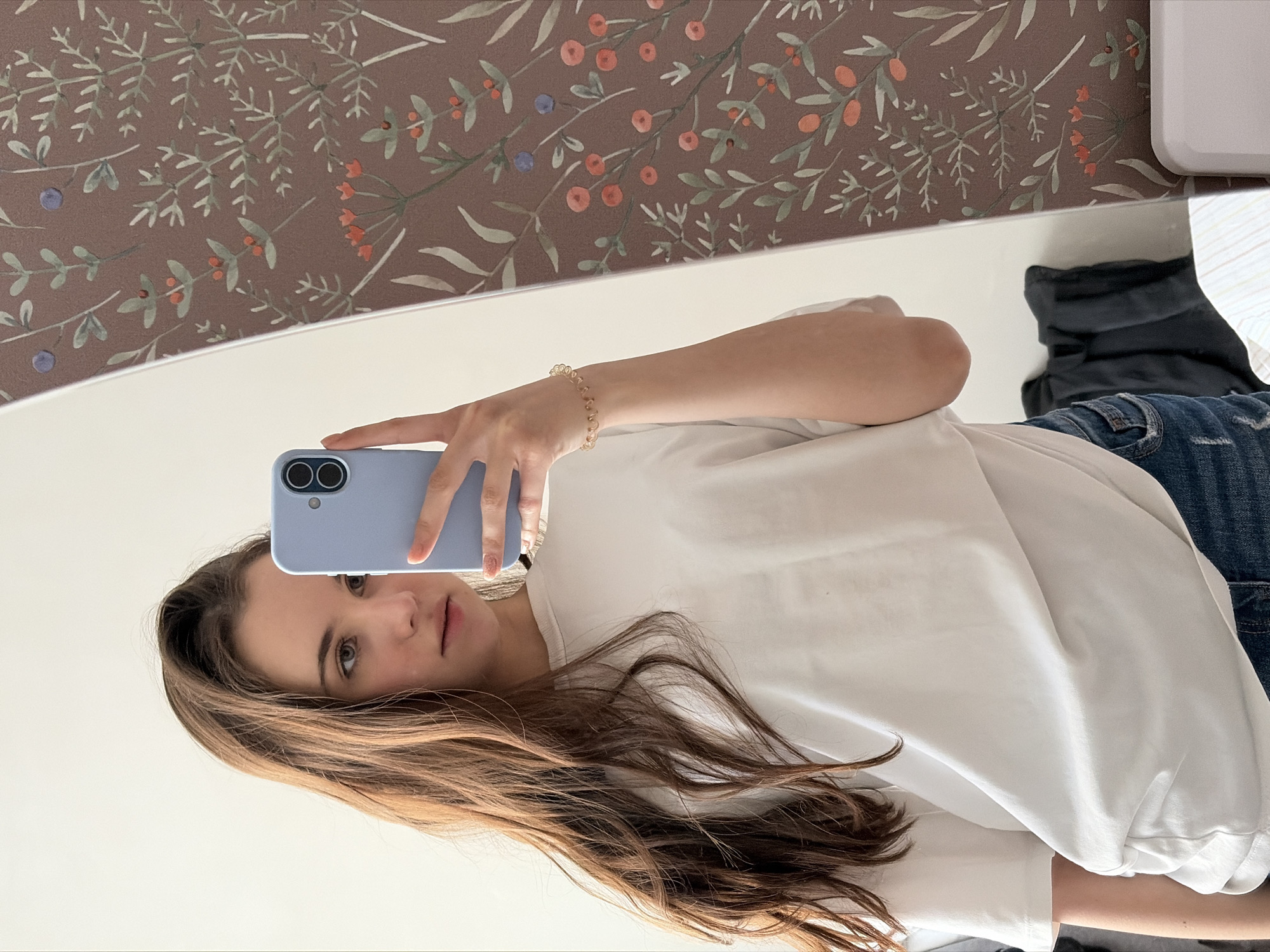 A mirrored reflection depicts a young woman holding a light blue smartphone up to take a selfie. She wears a white t-shirt and blue jeans, and a beaded bracelet is visible on her left wrist. Her hair is long and brown. Behind her is a wall covered in a floral pattern. A black jacket or similar clothing is seen partially in the reflection. The image does not provide any information about the location being Izhevsk, Russia.