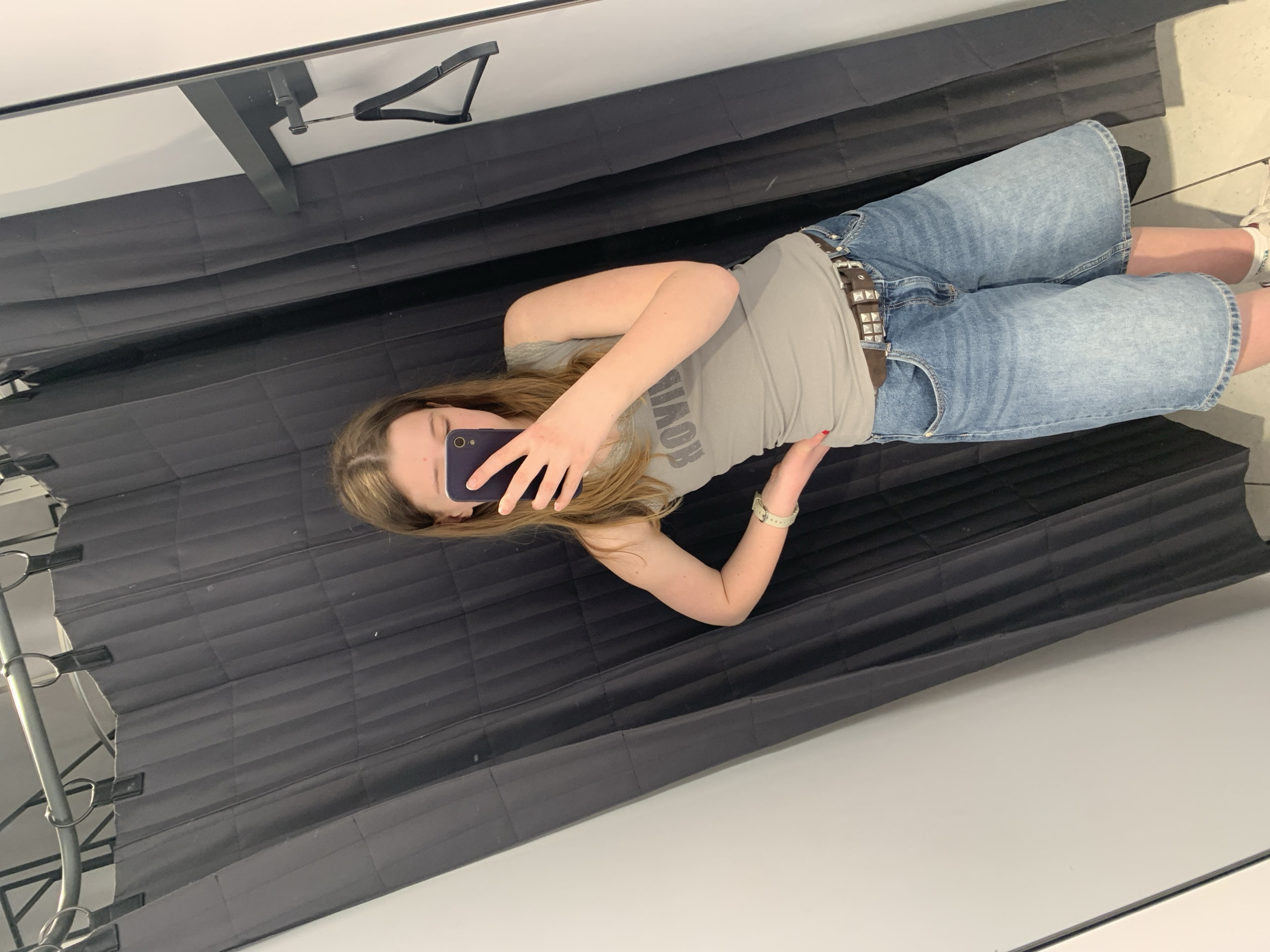 A young person is depicted taking a mirror selfie, appearing to be standing in what resembles a changing room. The image is oriented horizontally, showing the person rotated 90 degrees clockwise. The subject has long brown hair and is wearing a light grey, ribbed t-shirt that displays the word "BOYISH" in dark grey letters (visible in reverse in the mirror). They are dressed in light-wash denim Bermuda shorts, secured by a dark brown belt embellished with silver studs. A light-colored wristwatch is visible on their left wrist.
The person is holding a dark-colored smartphone with a purple-ish case in their right hand, using it to capture the photograph, which partially obscures their face. No specific emotions are discernible. The background of the scene behind the person consists of dark grey or black pleated fabric, suggesting a curtained-off area typical of a changing room or retail environment. Reflected in the upper portion of the mirror are elements resembling a black clothing hanger and parts of a metallic structure, possibly a clothing rack or fitting room frame. The setting indicates an indoor location, likely a retail store or boutique in Belgrade, Serbia, where the person is possibly trying on outfits.