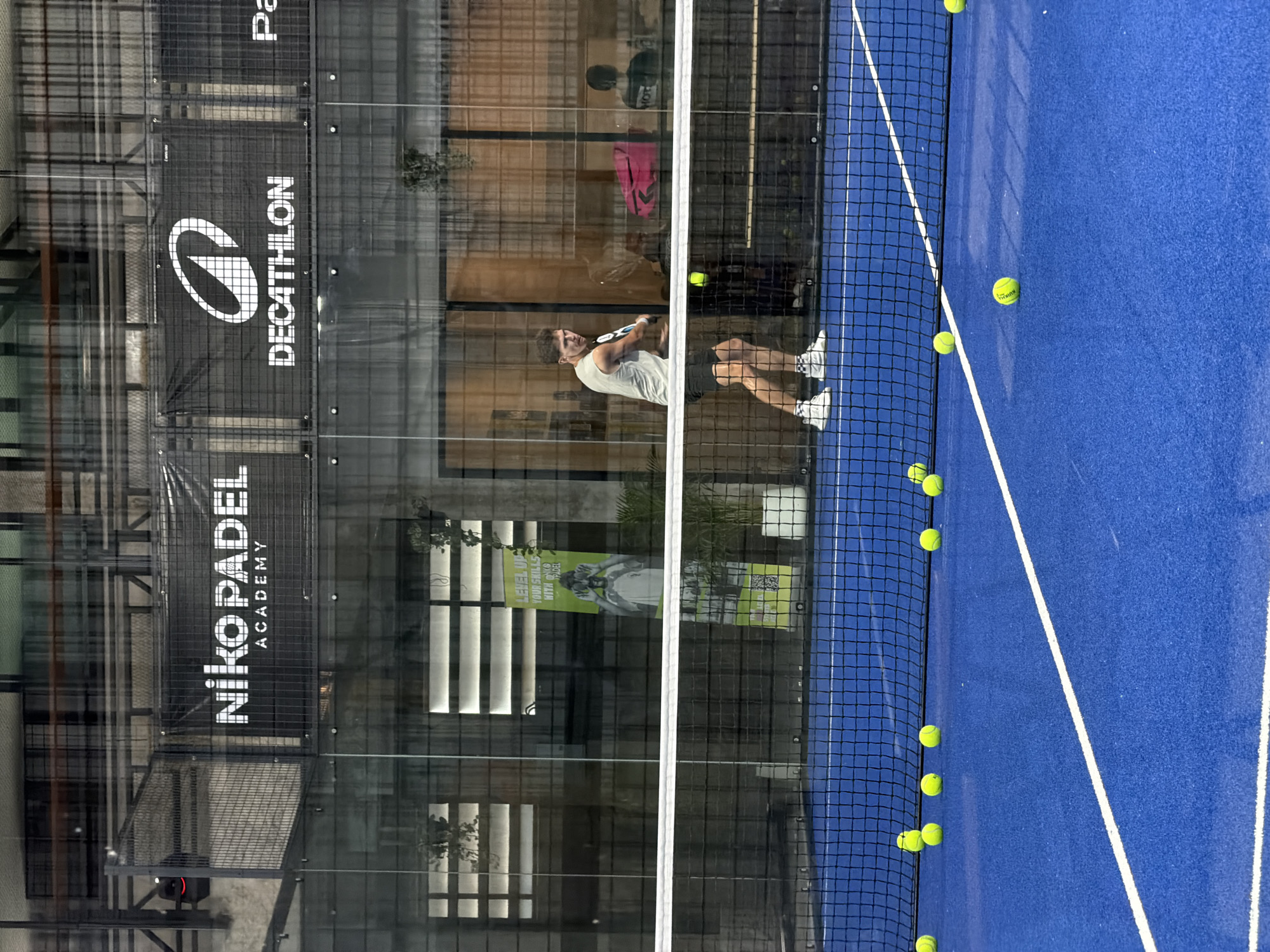 In Ban Buak Khrok North, Thailand, a young man in athletic attire is captured mid-swing on a vibrant blue padel court. He wears a light-colored sleeveless shirt, dark shorts, and white sneakers with black accents. The court is dotted with several yellow padel balls, some scattered near the net. A white net divides the court. Behind the net, the backdrop features glass walls with various advertisements and branding, including "Niko PADEL ACADEMY" and "DECATHLON". Indoor plants in white pots are visible, adding a touch of greenery to the setting. Reflections on the glass indicate an indoor environment.