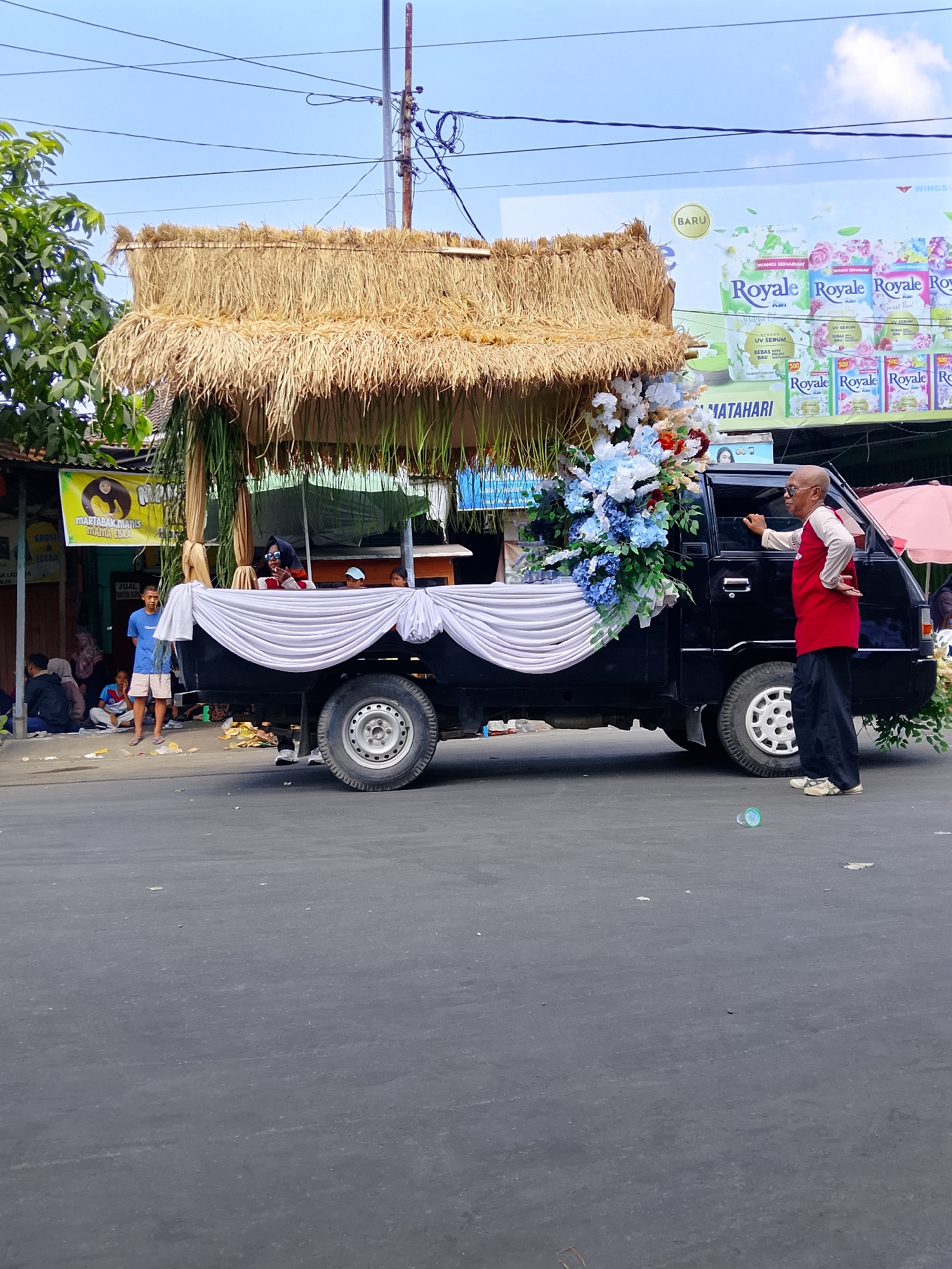 The image depicts a street scene in Balun, Indonesia, featuring a modified pickup truck acting as a float for a traditional event or parade. The truck's flatbed has been covered with white fabric draped in a decorative manner. On top of the flatbed, a thatched roof structure, reminiscent of a traditional hut or gazebo, has been constructed. This structure is adorned with a large, elaborate floral arrangement primarily composed of white and blue flowers, with some red accents. Green leafy branches also hang from the roof.
In the foreground, to the right of the truck, a bald, elderly man with glasses is standing. He is wearing a red long-sleeved shirt with white sleeves and dark pants, and appears to be gesturing with his right hand towards the back of the truck. His left hand is behind his back.
On the left side of the truck, a young boy in a blue t-shirt and light-colored shorts stands on the side of the road. Several other people are seated or standing on the sidewalk to the left, some appearing to be onlookers or participants in the event. A banner with a yellow circular logo and text is visible on a building behind the people.
The environment suggests a bright, sunny day with a clear blue sky. Overhead, power lines and a utility pole are visible. In the background, a large billboard for "Royale" fabric softener is prominently displayed, featuring images of flowers and product packaging. A pink umbrella is partially visible behind the truck. The road surface appears to be asphalt, with some scattered debris. The scene captures a moment of local cultural activity.