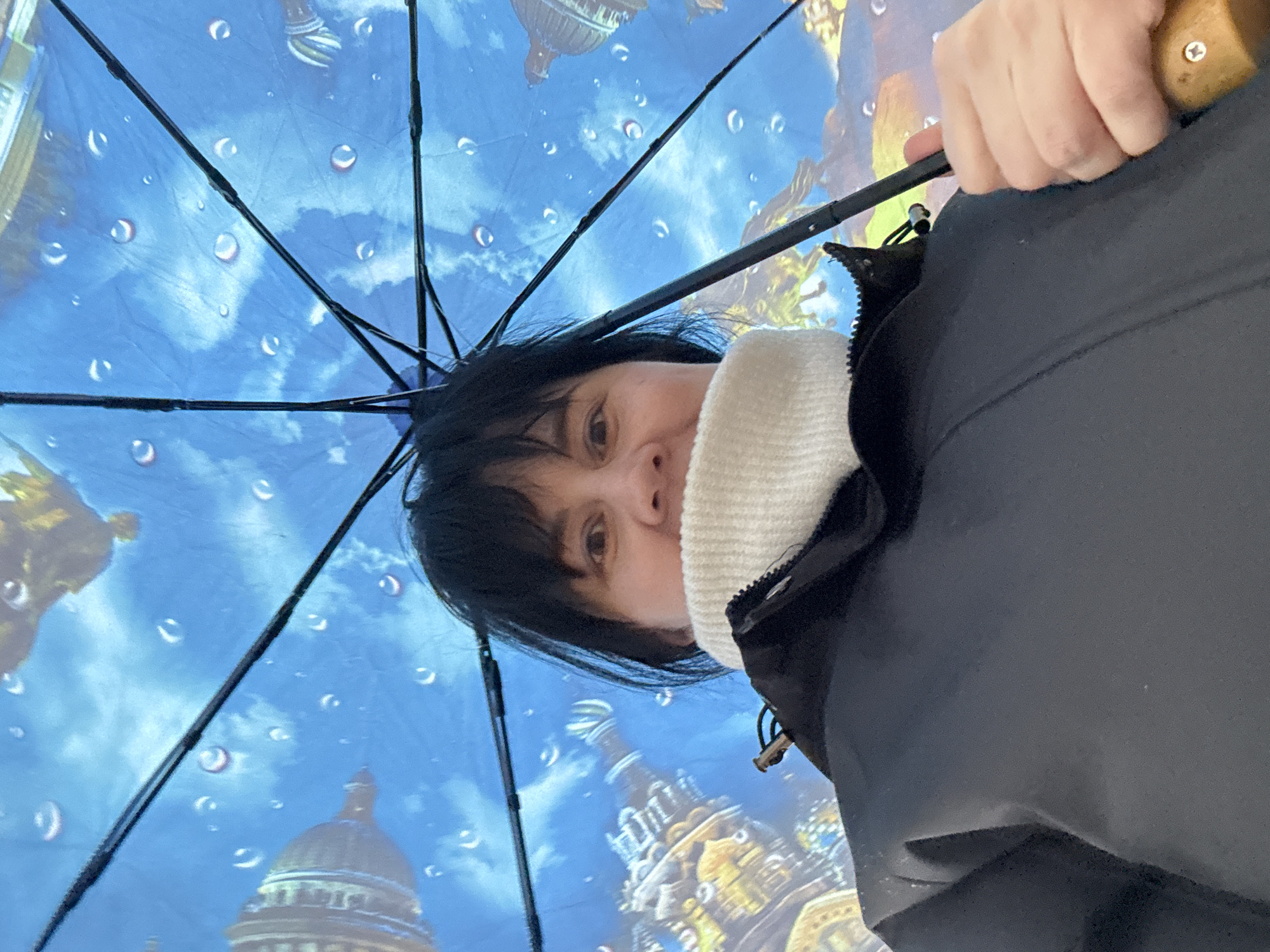 A woman stands beneath an umbrella with a blue background and images of buildings and raindrops. The umbrella's spokes are black. She is wearing a black coat and a white sweater. The scene is not in Zav'yalovo, Russia, as buildings like that are not there.