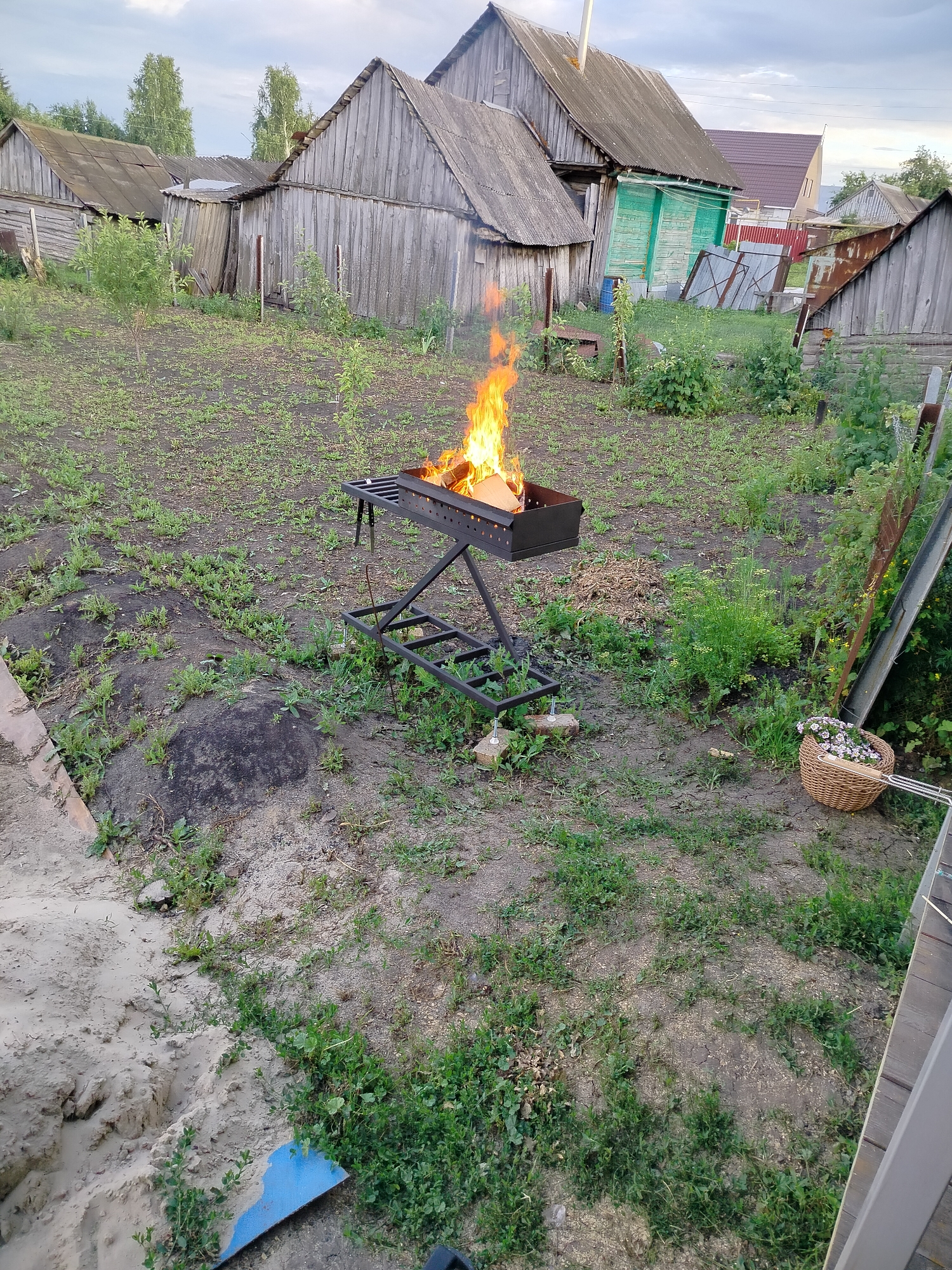 The image depicts a rural outdoor scene in the village of Vazery, Russia. In the foreground, a metal brazier is alight with a fire, with pieces of wood visible within. The brazier is situated on a grassy, uneven area with patches of bare earth. To the right of the brazier, a woven basket filled with white flowers sits on the ground, next to a metal pole. Further into the background, several weathered wooden structures, likely sheds or outbuildings, are visible. A house with a red roof and green siding is also present. The sky is overcast with a hint of clouds. The ground is covered with grass and dirt, with some sand visible in the lower left corner.